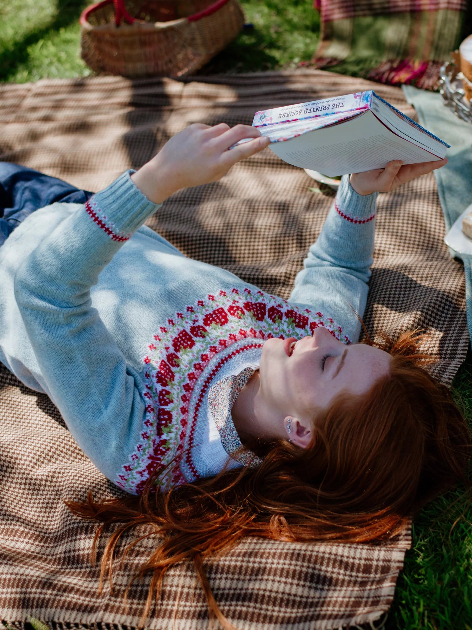 A person with long red hair relaxes outdoors on the Campbells of Beauly Crofters Blanket, reading a book in a light blue sweater with red and white patterns, surrounded by picnic items for a classic country look.