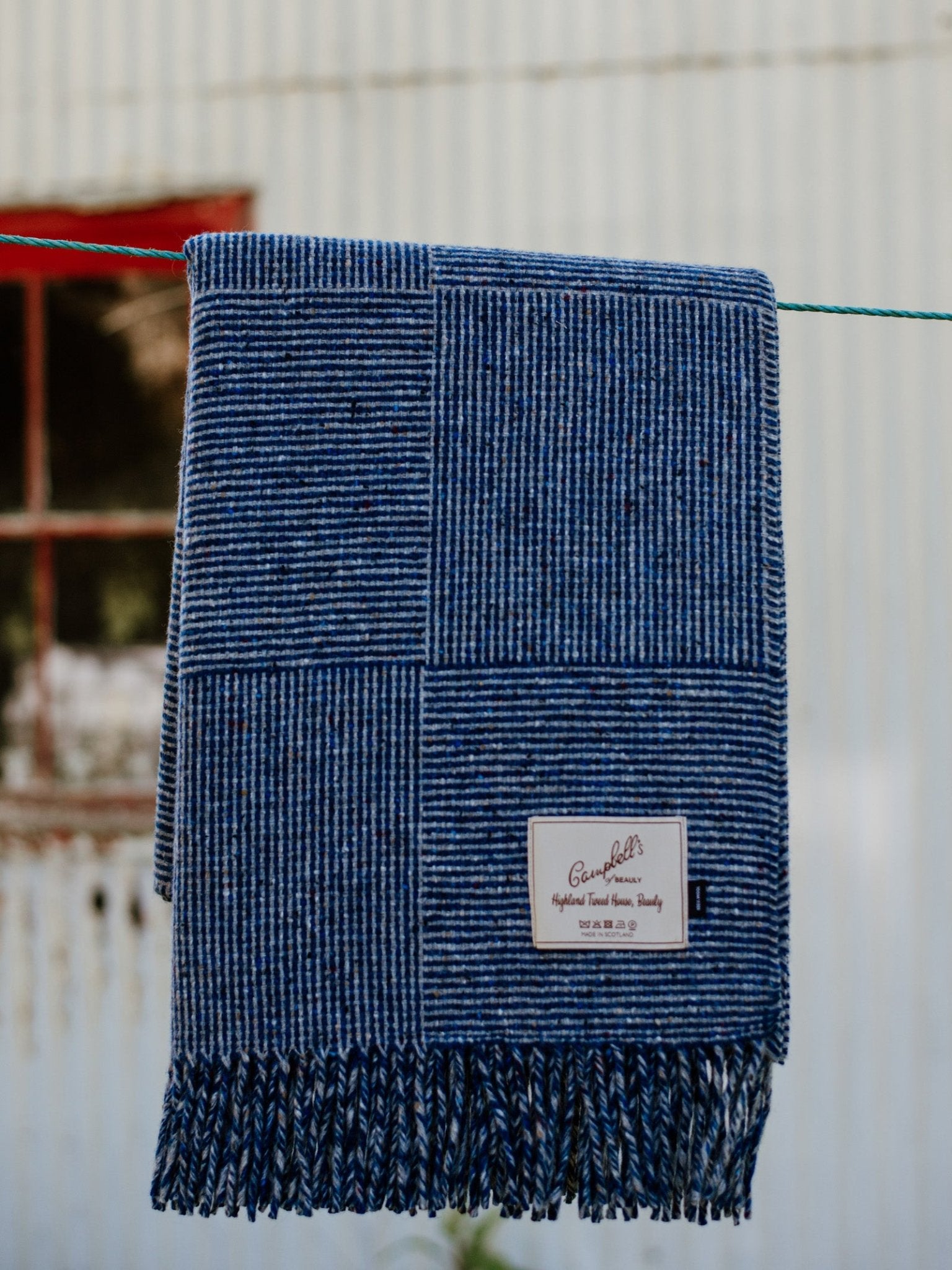The Cabrich Lambswool Throw by Campbells of Beauly, featuring a blue patchwork stripe design and fringe, hangs on a clothesline. A white label is stitched in the lower right corner with a blurred rustic window and corrugated wall behind.