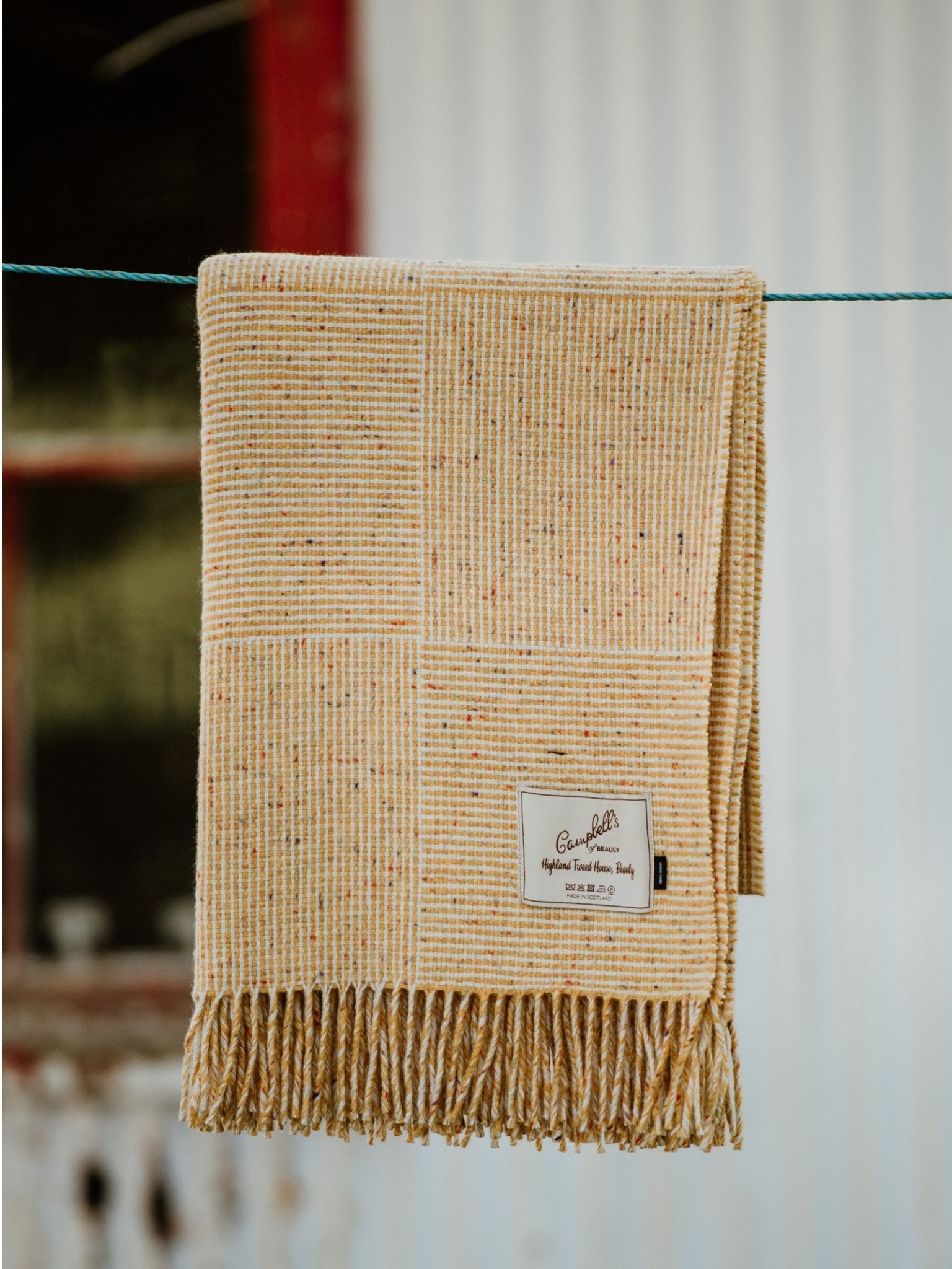 The Cabrich Lambswool Throw by Campbells of Beauly, featuring a fringed edge and fabric label, is draped over a green clothesline. Its yellow basket weave design contrasts with the blurred window and corrugated metal wall behind.