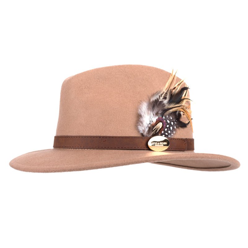 The Hicks & Brown Gamebird Fedora in camel is crafted from wool felt and features a dark brown band with a feather wrap plus a small gold oval pin on the side.
