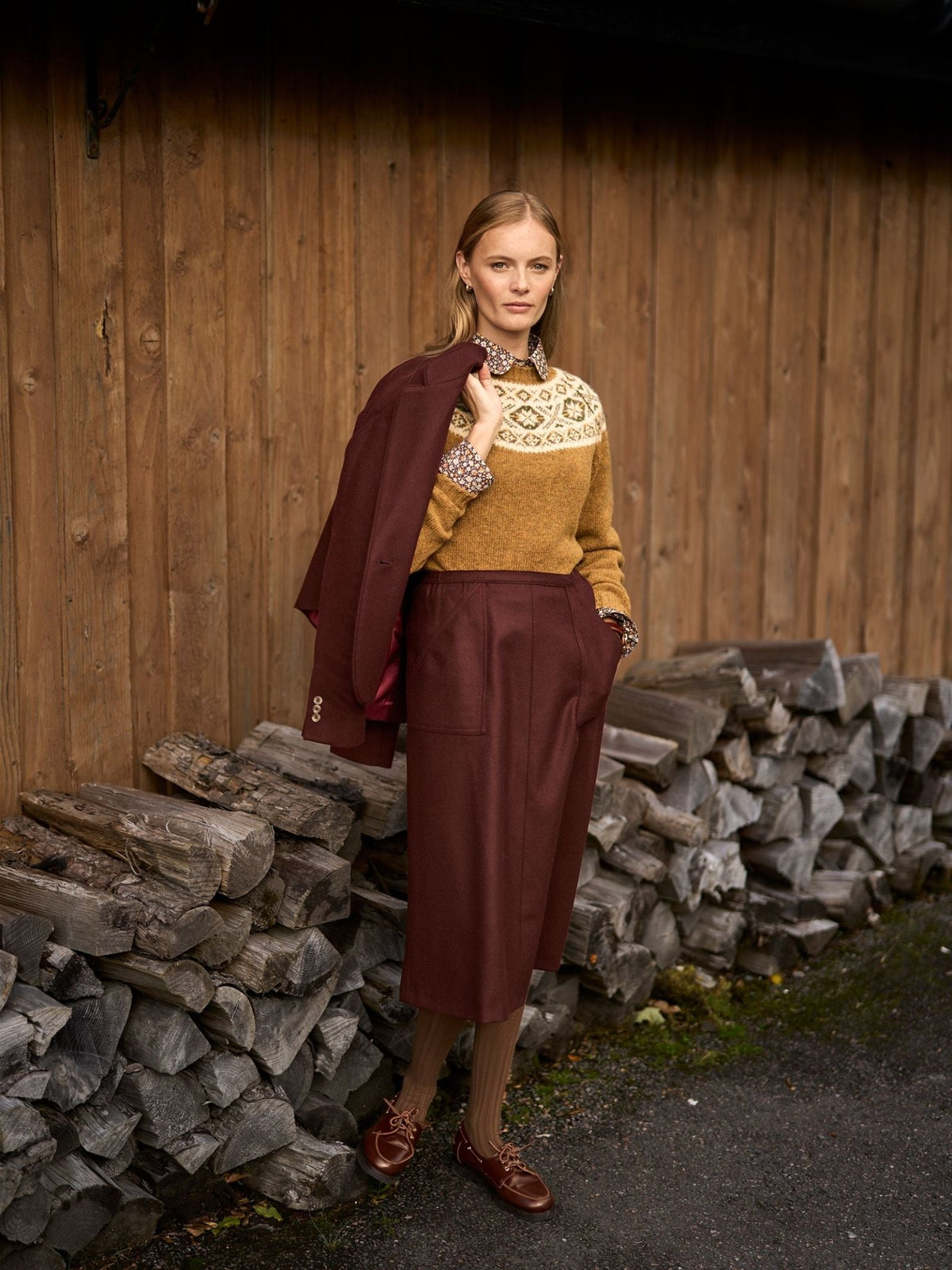 A woman wears a mustard yellow patterned sweater and a dark red British tweed A-Line Skirt by Campbell’s of Beauly, holding a matching jacket over her shoulder while standing in front of a woodpile.