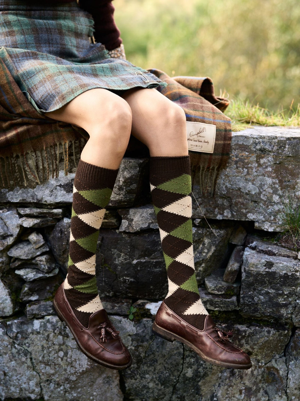 A person sits on a stone wall wearing Campbell’s of Beauly Argyle Socks in shades of brown, green, and cream, paired with brown leather tassel loafers.