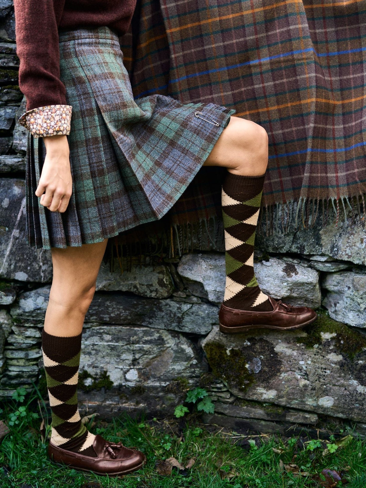Person wearing a plaid kilt and Campbell’s of Beauly Argyle Socks with knee-high patterns and brown leather loafers, standing by a stone wall.