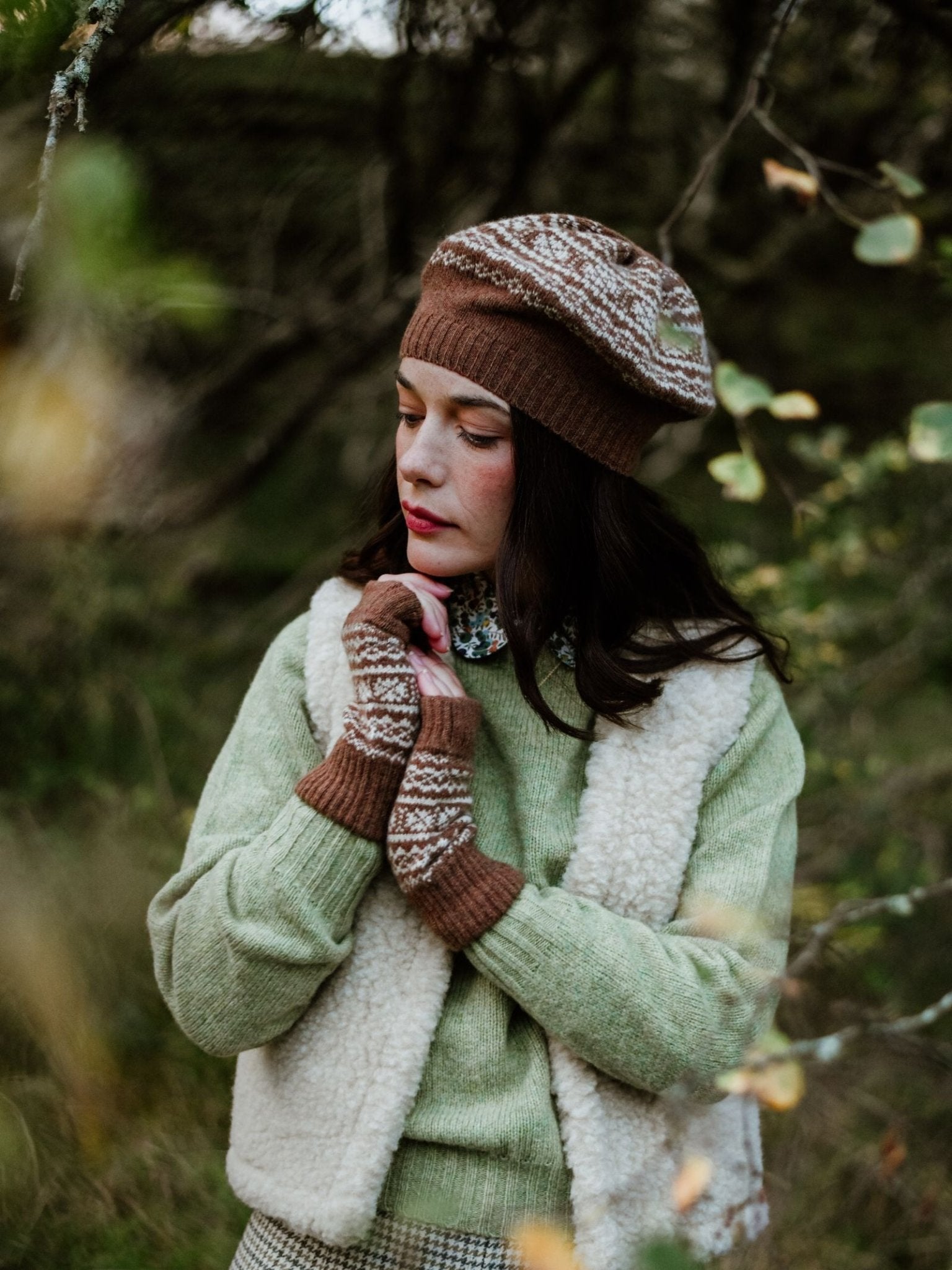A woman stands outdoors among trees, looking down thoughtfully, wearing the Auld Stock Two Colour Fairisle Beret in Chocolate by Campbell’s of Beauly. Her cozy ensemble—a brown patterned beret paired with fingerless gloves, a green sweater, and a white fleece vest—evokes the timeless charm of Scottish knitwear.