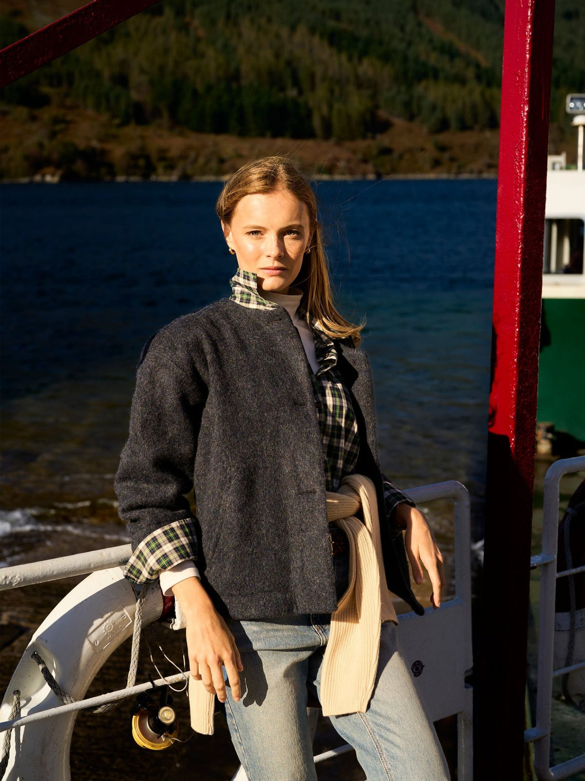 A person wearing a timeless dark Brushed Wool Jacket by Campbell’s of Beauly with plaid cuffs, standing on the deck of a boat by a lake.