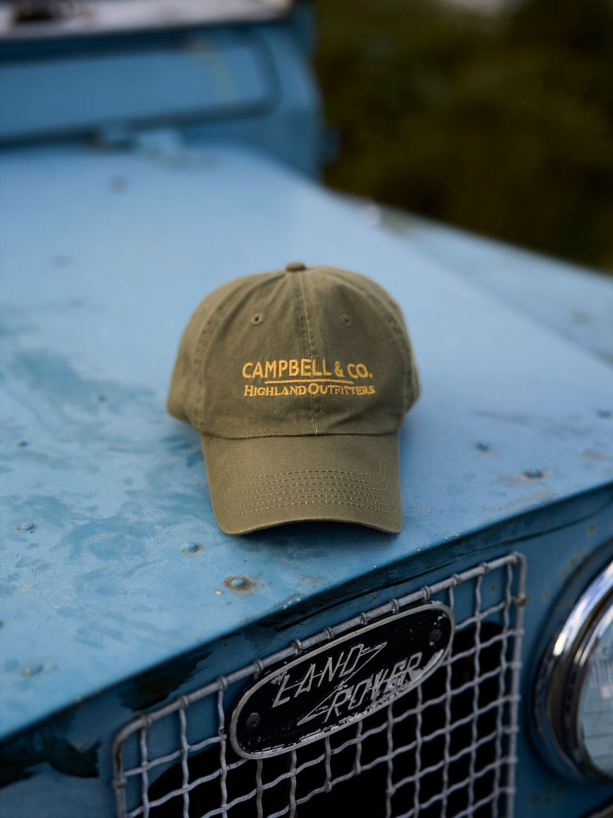 An olive green Campbell’s Baseball Cap with yellow embroidery reading CAMPBELL & CO. HIGHLAND OUTFITTERS resting on the hood of a blue vintage vehicle.