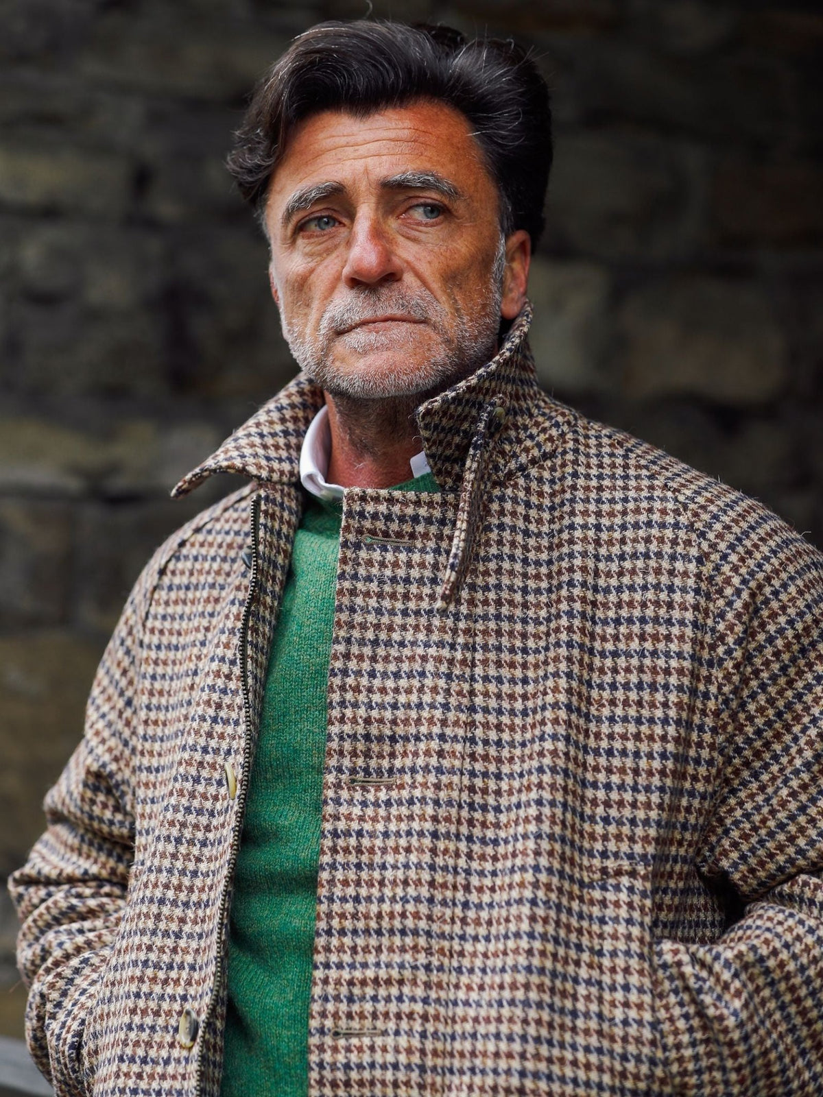 A man with gray hair and a beard stands outdoors in front of a stone wall, looking thoughtfully to the side while wearing the Cannich Coat from Campbell’s of Beauly layered over a green sweater—perfectly embodying the timeless elegance of country pursuits.
