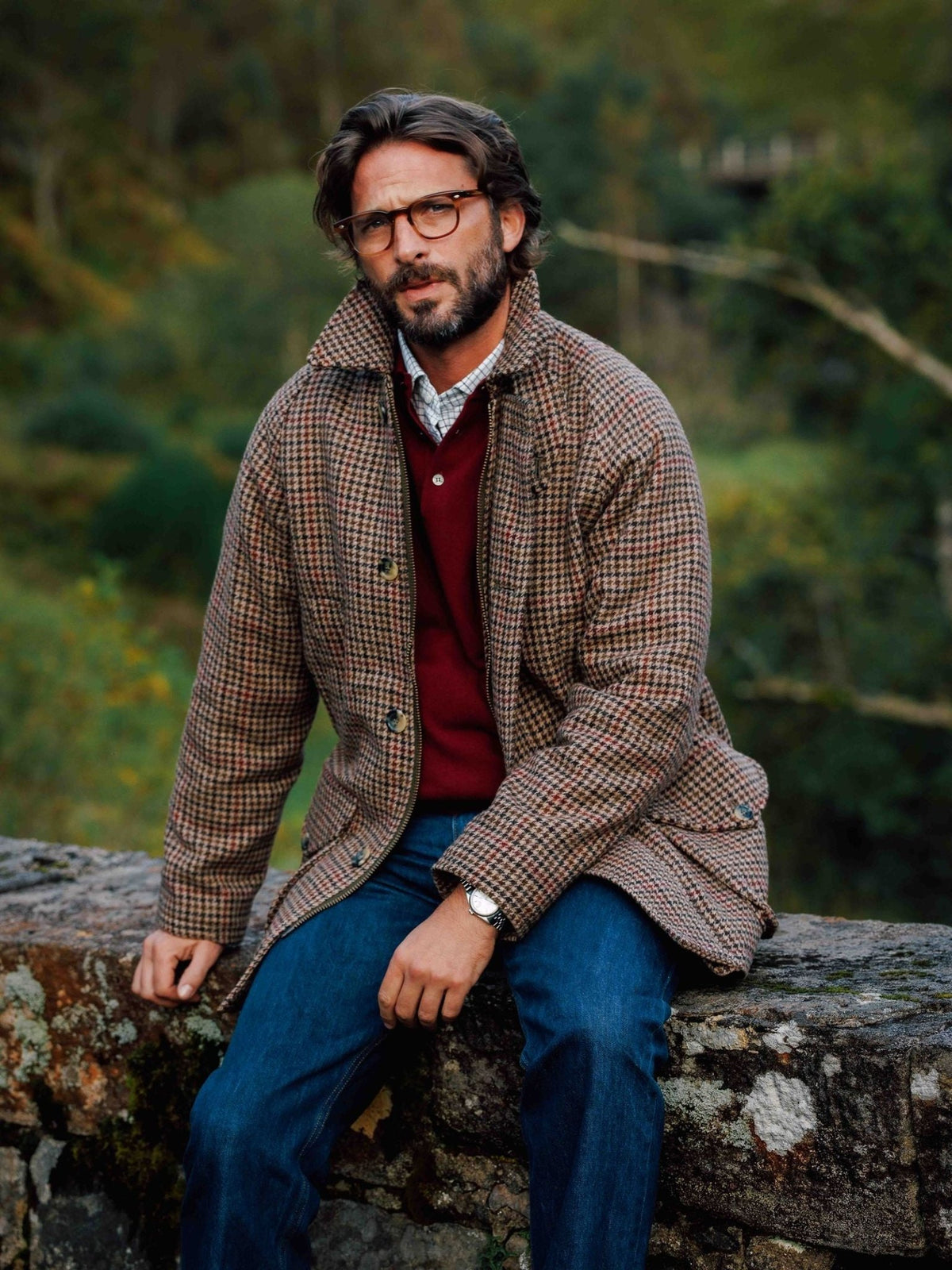 A man wearing a brown patterned Campbell’s of Beauly Cannich Coat over a red sweater and collared shirt sits on a stone wall.