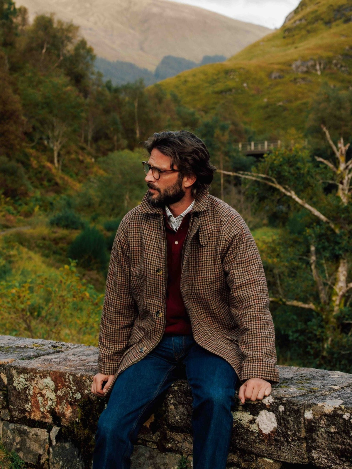 A man with a beard and glasses sits on a stone wall wearing a brown houndstooth Cannich Coat by Campbell’s of Beauly over a maroon sweater and blue jeans.