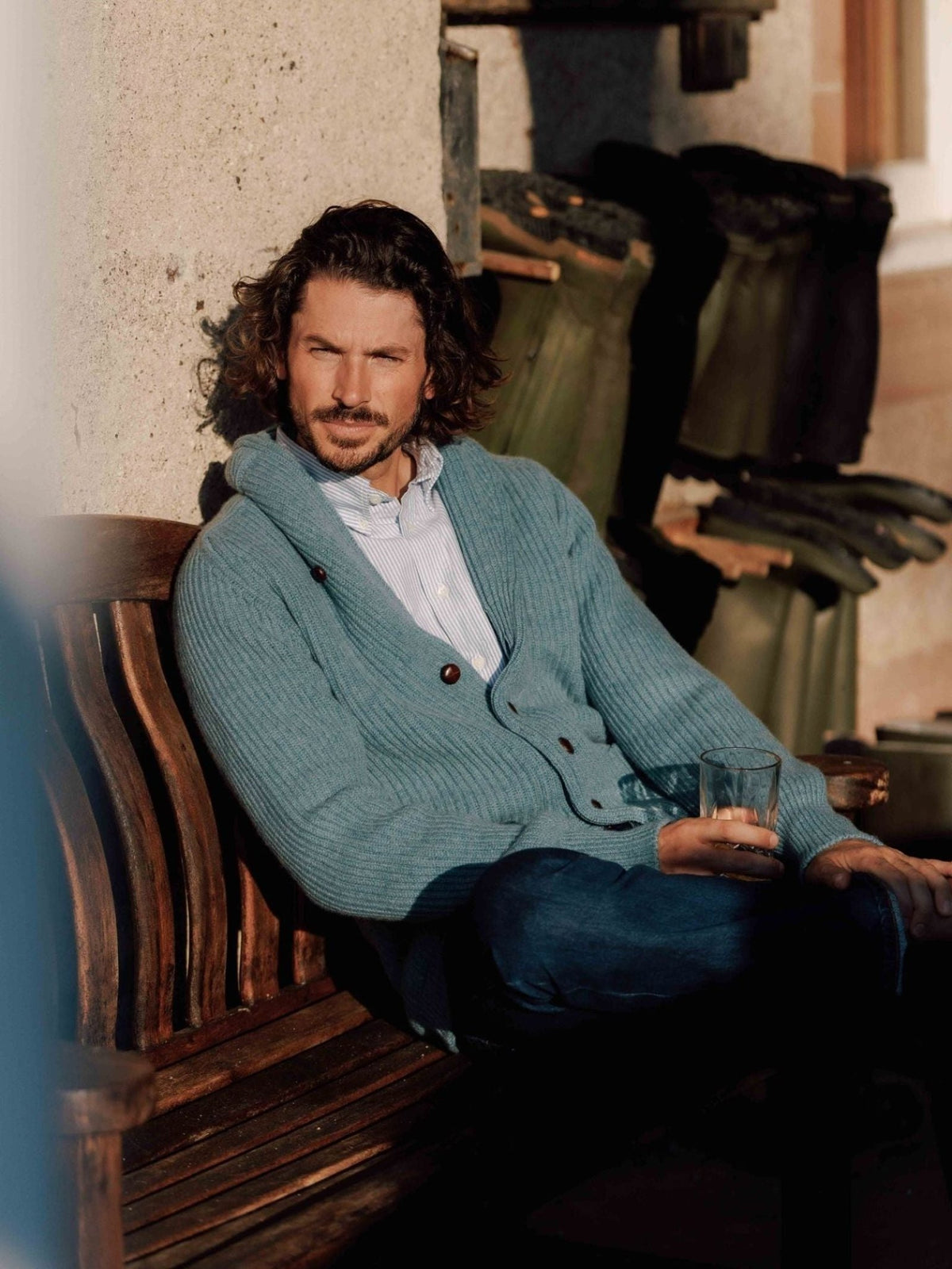 A man with wavy hair, dressed in a Campbell’s of Beauly Cashmere Shawl Collar Cardigan and jeans, sits on a wooden bench holding a glass. Sunlight illuminates him while trash bins appear in the background.