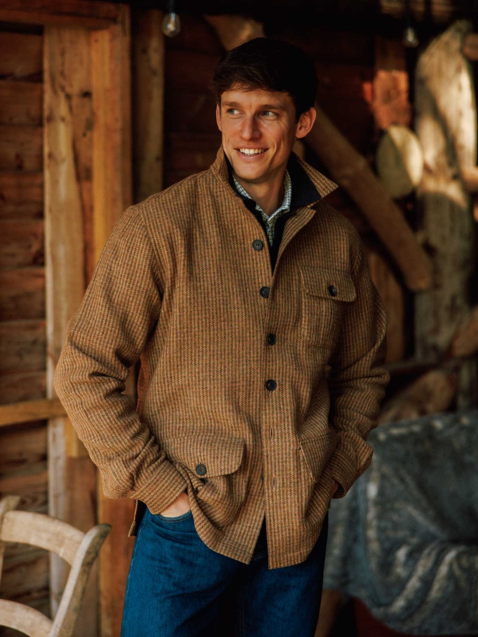 A man stands indoors with his hands in his pockets, smiling and looking to the side, dressed in Campbell’s of Beauly’s Casual Shacket layered over a plaid shirt. The brown checkered jacket, inspired by classic British tweed, is complemented by a rustic wooden background with a chair and blanket that underscores the relaxed tailoring of his casual look.