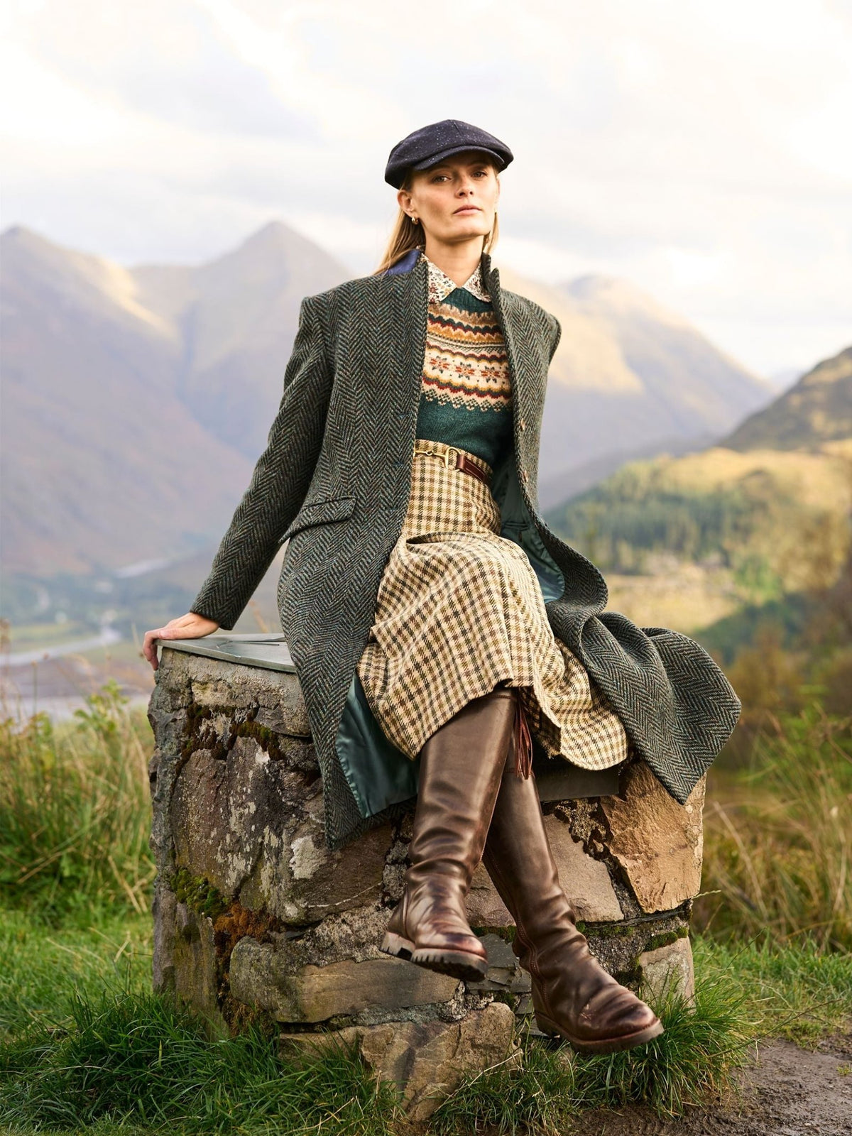 A woman wearing a Donegal Herringbone Coat, patterned sweater, plaid skirt, and tall leather boots sits on a stone wall in a mountainous landscape.