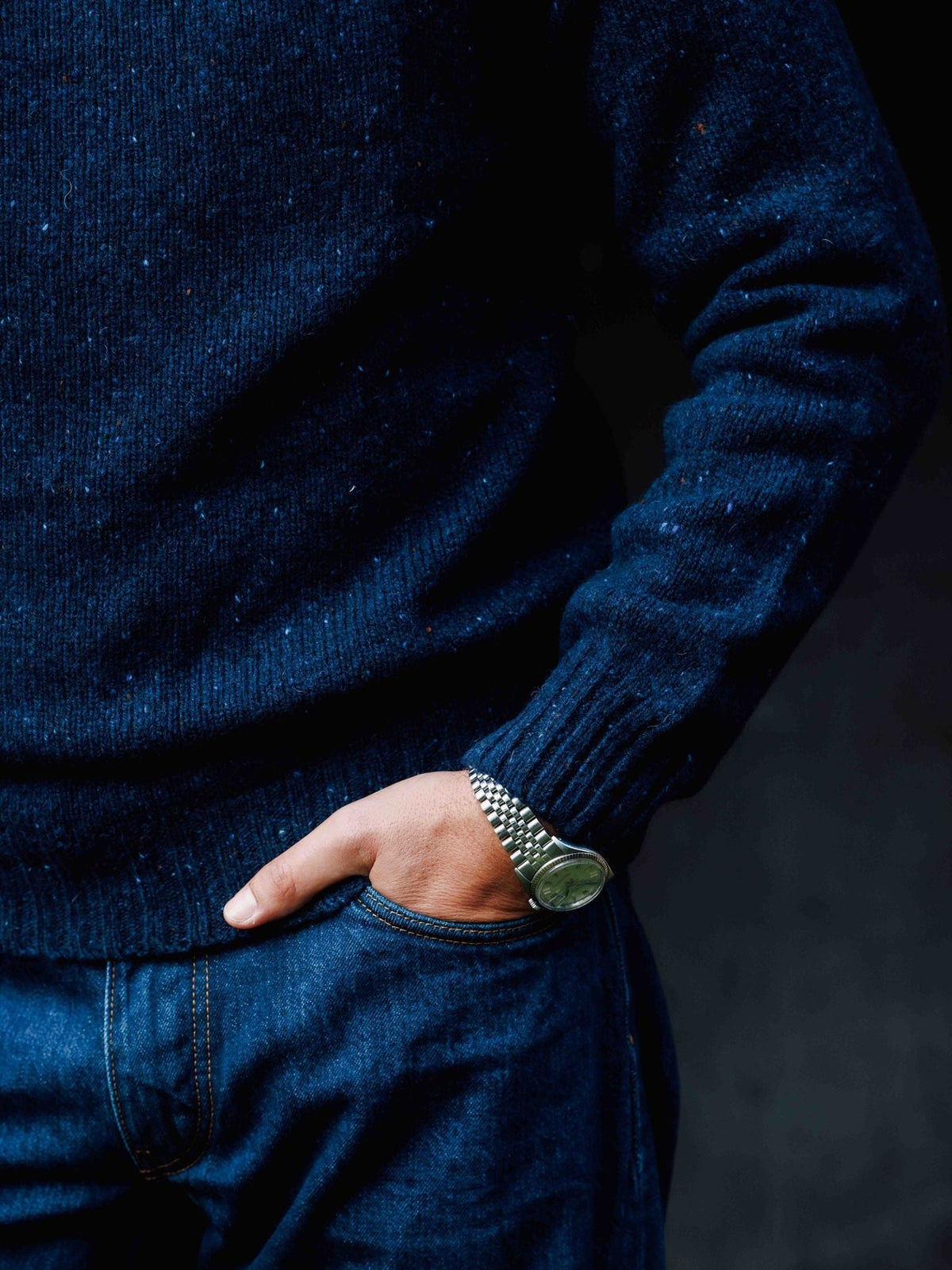 A person wearing a navy blue Donegal Lambswool Crew Neck Jumper and blue jeans, with a metallic wristwatch visible on their wrist.