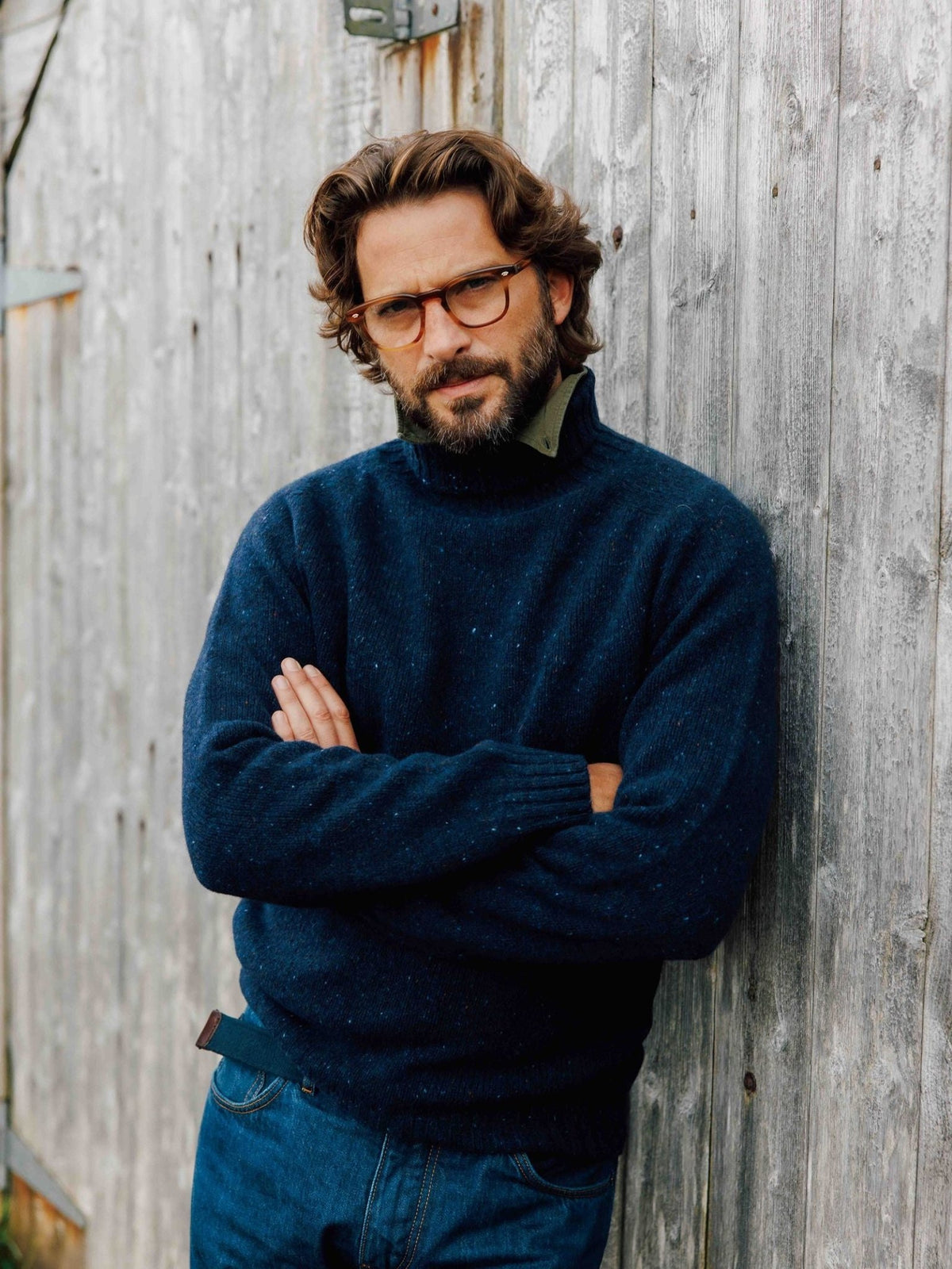 A man wearing a dark blue Donegal Lambswool Roll Neck sweater with a dark green shirt underneath, leaning against a wooden wall.