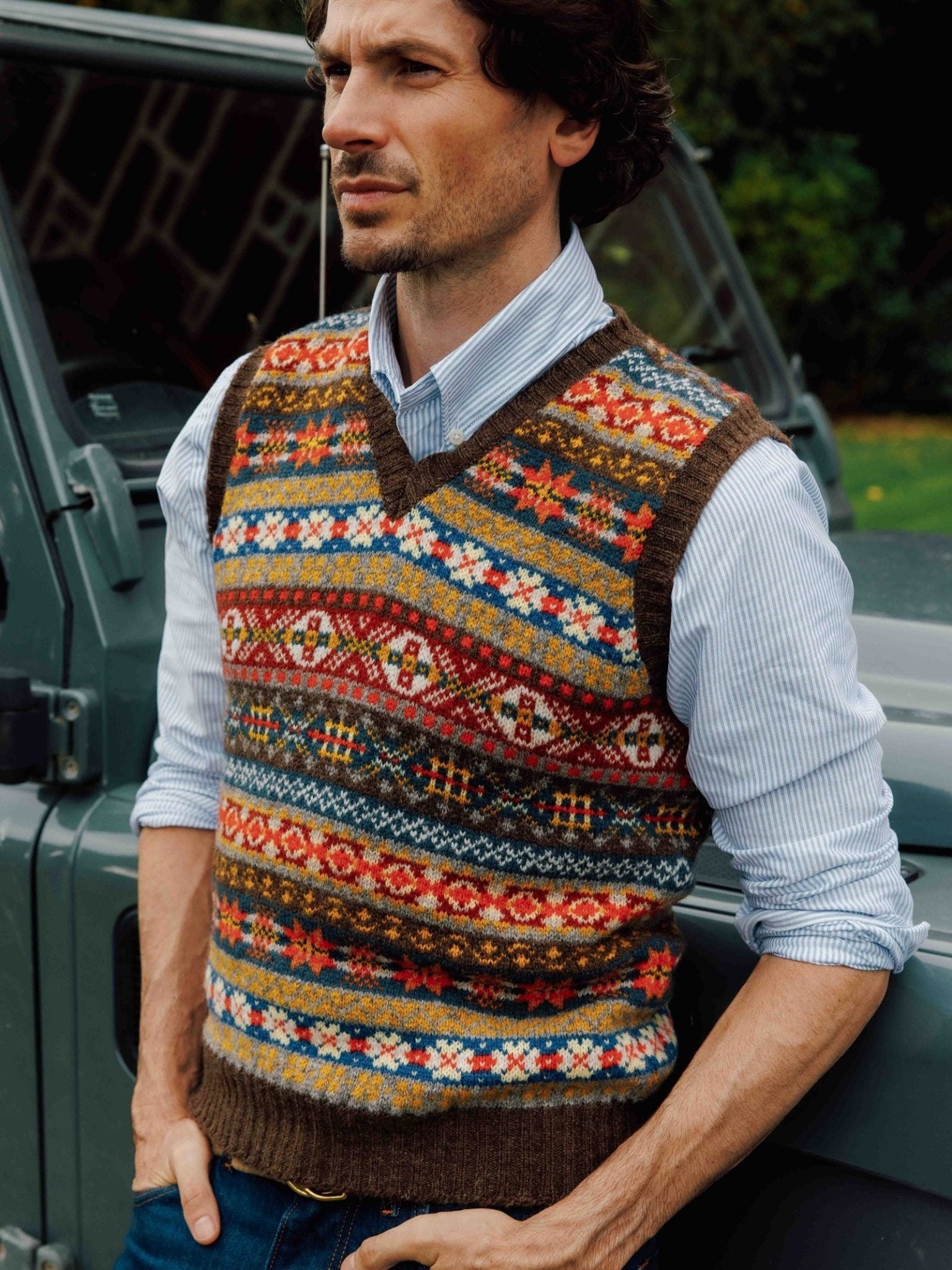 A man wearing the Campbell’s of Beauly Fairisle Slipover—striped and colorful, knitted from warm Shetland wool—as a sleeveless layer over a light blue shirt stands beside a gray vehicle, gazing thoughtfully into the distance.