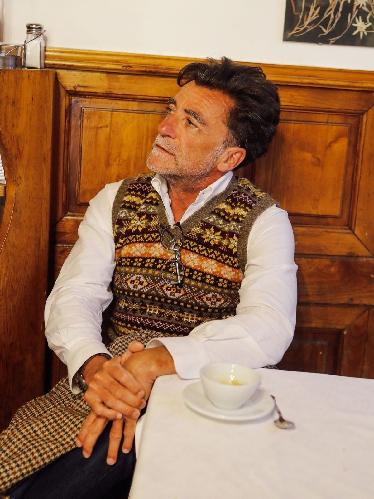 A man with dark hair and a beard, wearing the Campbell’s of Beauly Fairisle Slipover crafted from Shetland wool over a white shirt, sits at a table with a white cup, looking thoughtful; wooden paneling and artwork can be seen in the background.