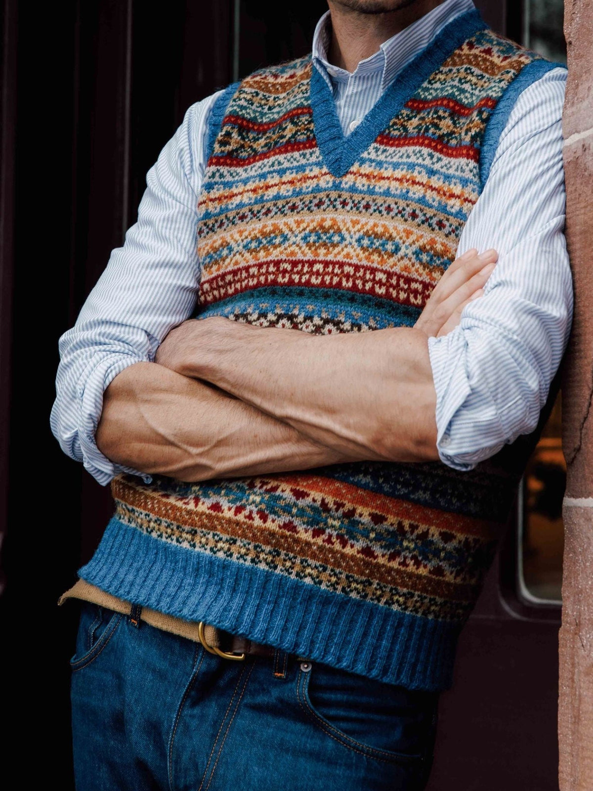 A man wearing the Campbell’s of Beauly Fairisle Slipover, crafted from Shetland wool, over a striped shirt and blue jeans stands with his arms crossed in front of a brown door.