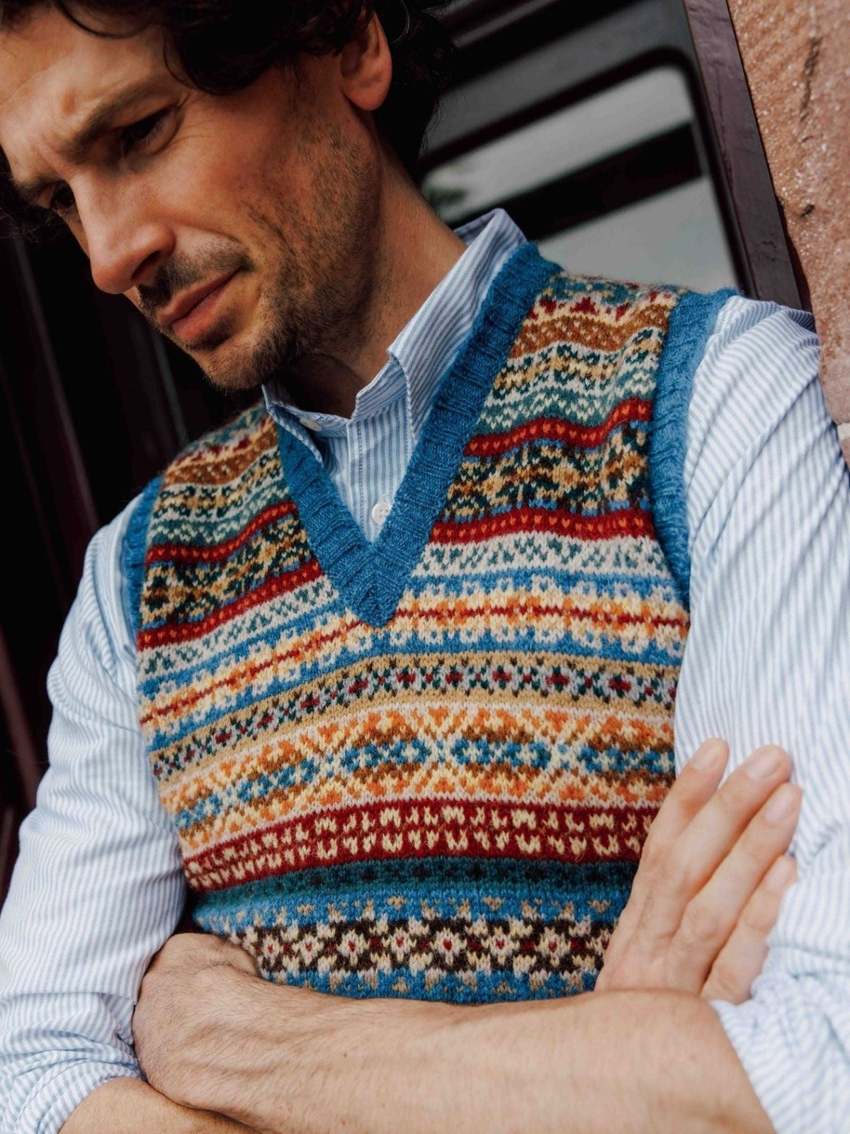 A man wearing the Campbell’s of Beauly Fairisle Slipover, crafted from patterned Shetland wool, stands with arms crossed over a striped shirt, gazing down thoughtfully.