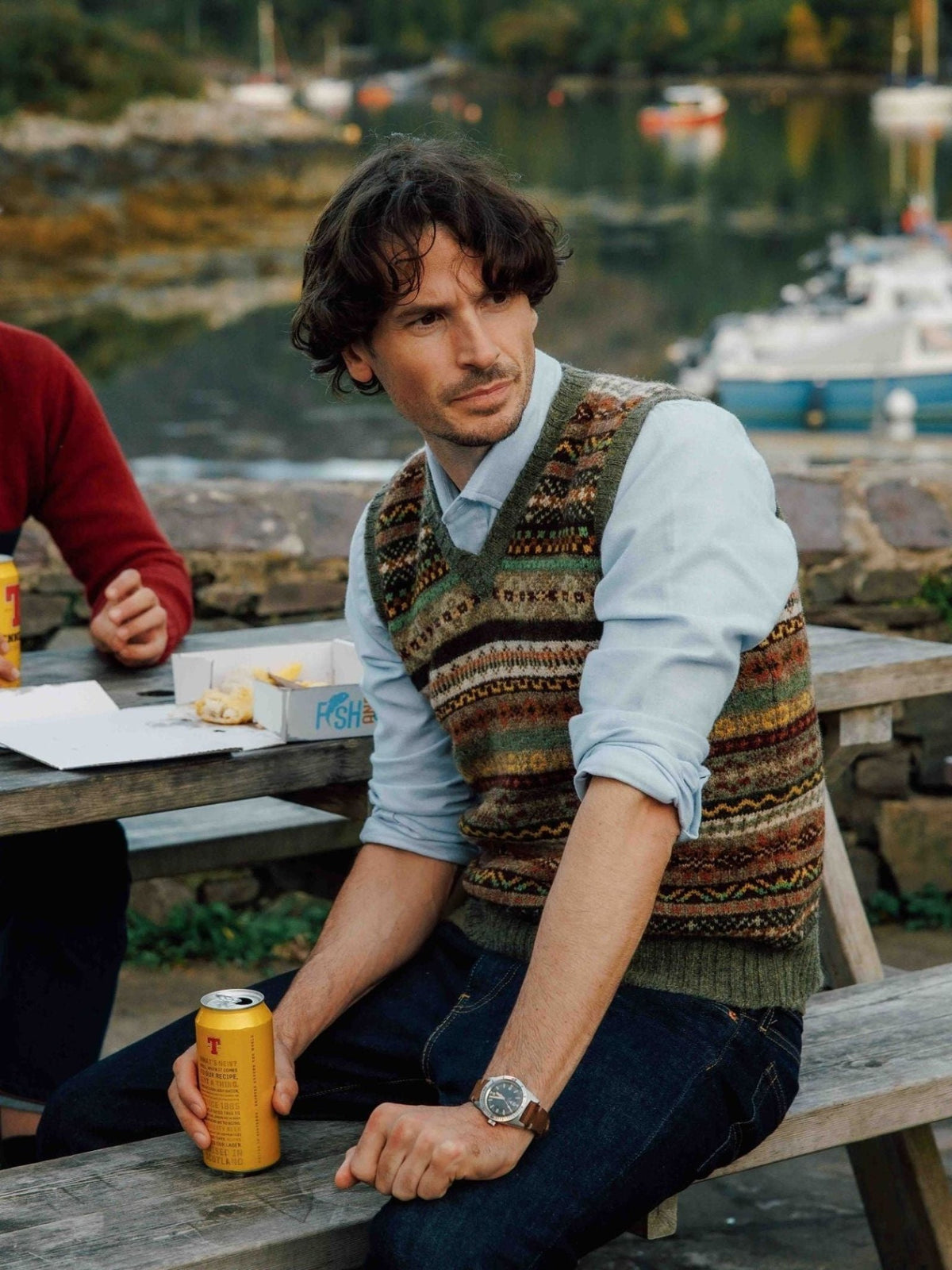 A man with wavy hair is seated on a wooden bench outdoors, holding a can and wearing the Campbell’s of Beauly Fairisle Slipover. Water, boats, and greenery can be seen in the background.