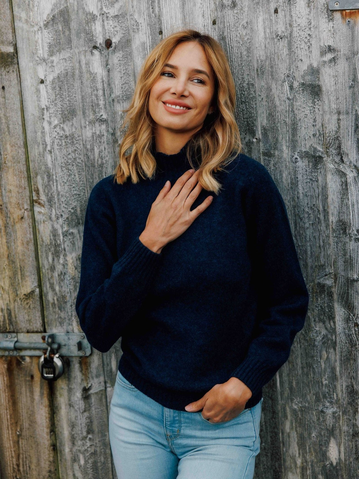A smiling woman wearing a dark blue Campbell’s of Beauly Geelong Lambswool Funnel Neck sweater with light wash jeans.