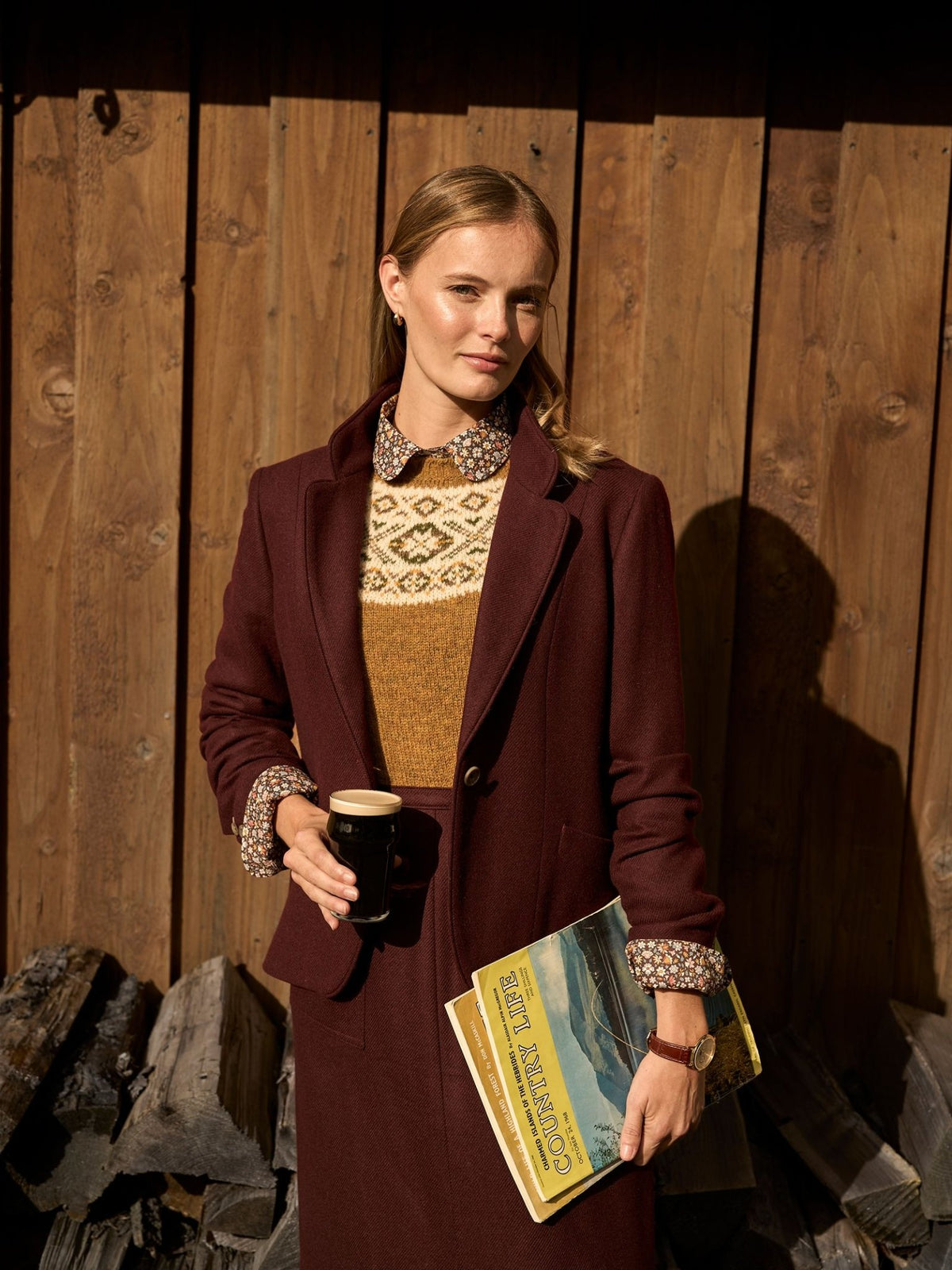 A woman wearing a gold herringbone House Jacket by Campbell’s of Beauly over a patterned shirt and a fair isle sweater, holding a glass of dark beer and a vintage magazine.