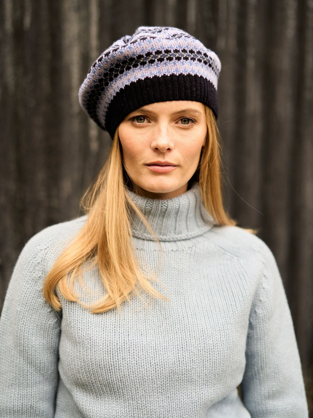 A woman wearing a light blue turtleneck sweater and a Lambswool Fairisle Beret in shades of purple and black.