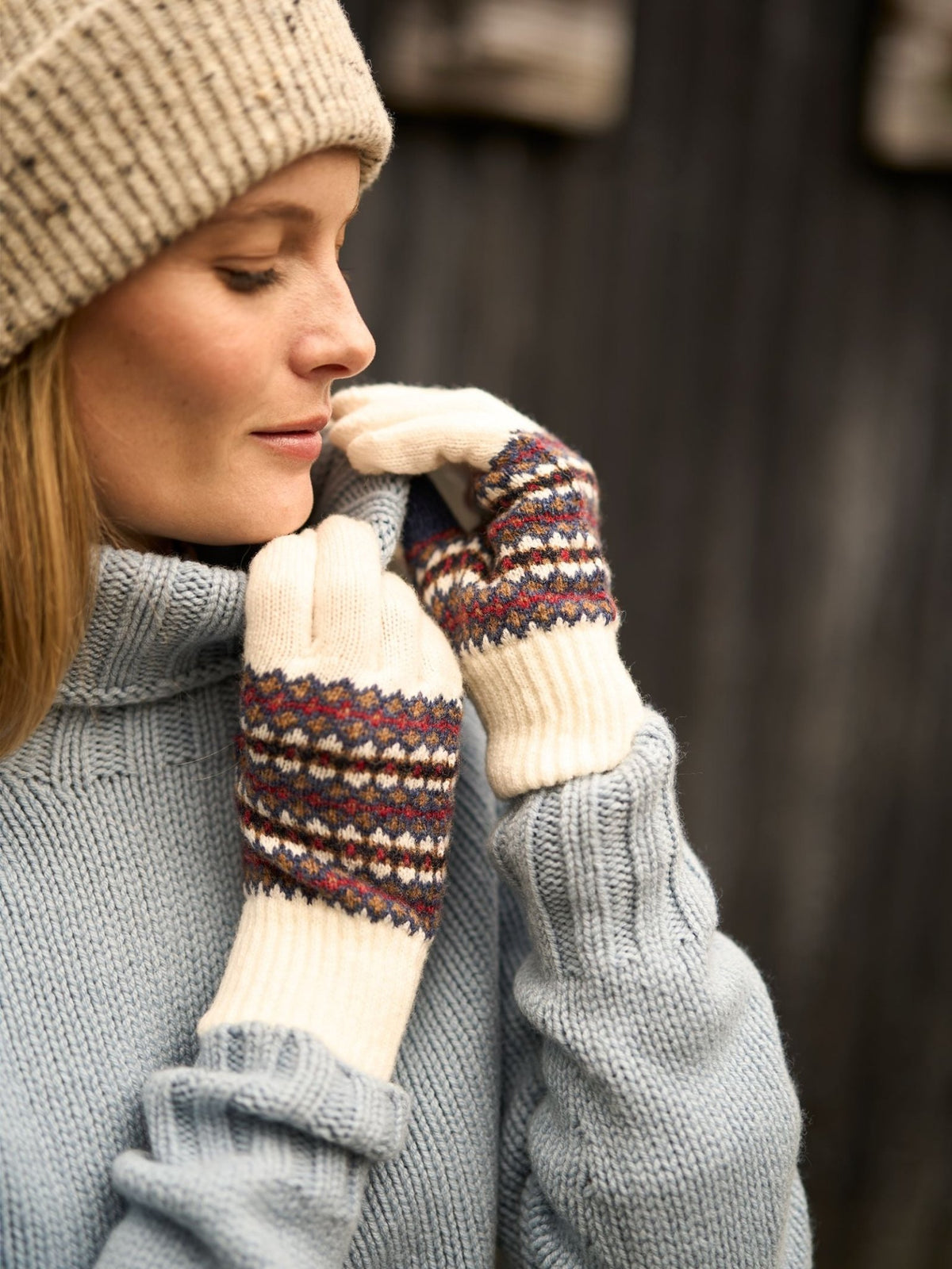 A person wears a knit beanie and blue turtleneck sweater, with their hands tucked into Campbell’s of Beauly Lambswool Fairisle Gloves.