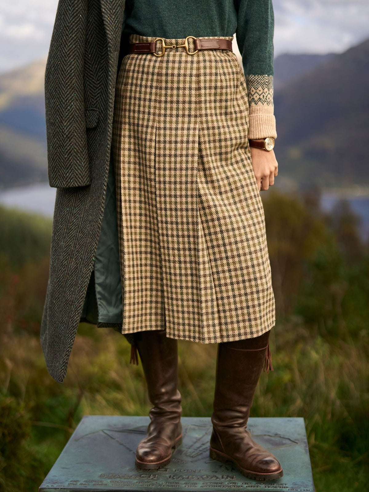 A person wearing a pleated plaid skirt, a green patterned sweater, a long tweed coat, and brown knee-high Leather Spanish Boots by Campbell’s of Beauly.