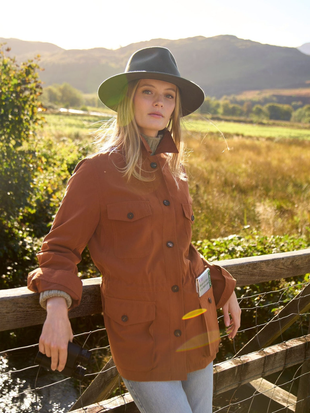 A person wearing a brown Lightweight Aigas Field Jacket by Campbell’s of Beauly and a dark hat leans against a wooden fence in a field, holding a pair of binoculars.