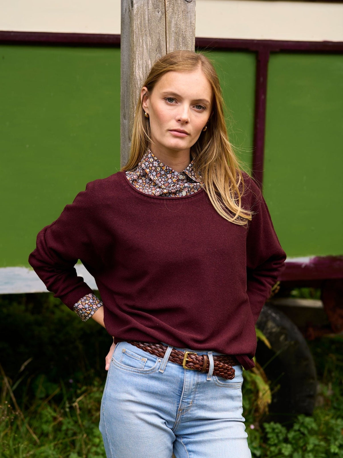 A person wearing a burgundy Merino Cashmere Boat Neck Jumper over a floral print collared shirt and light-wash jeans with a braided leather belt.