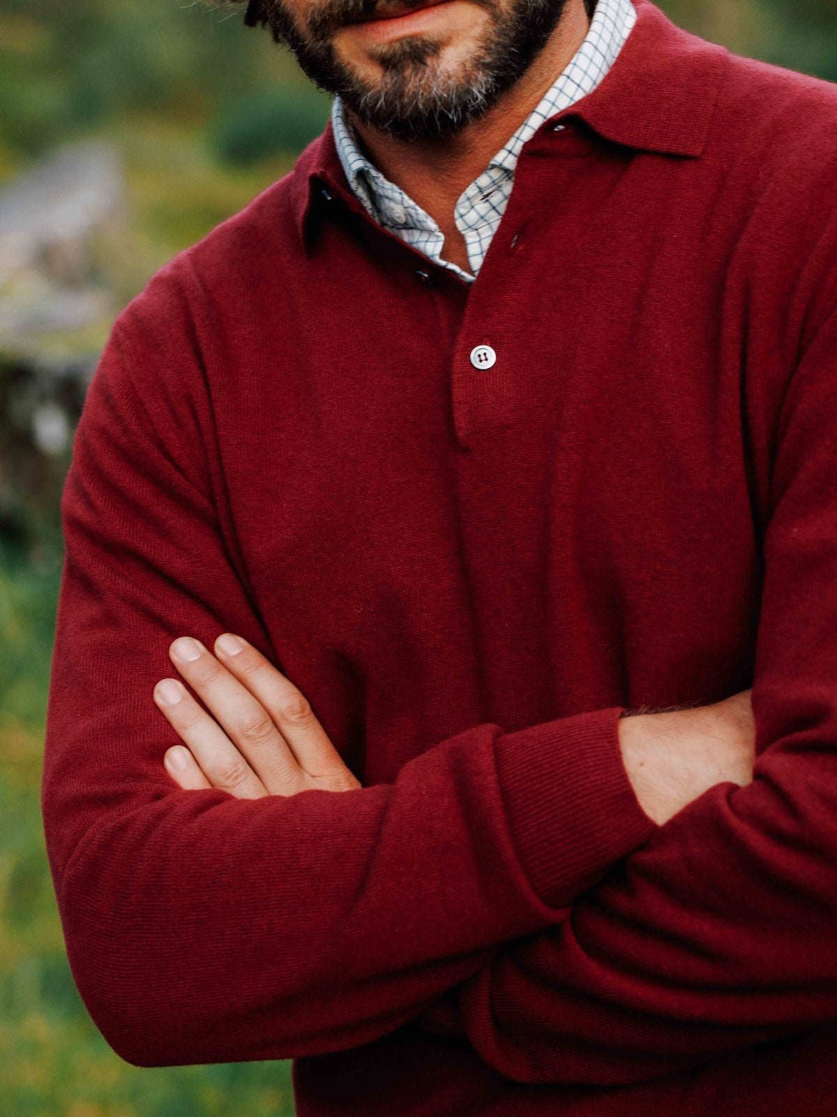 A man with his arms crossed wears a burgundy Merino & Cashmere Rugby by Campbell’s of Beauly over a plaid collared shirt, showcasing elegant Italian knitwear.