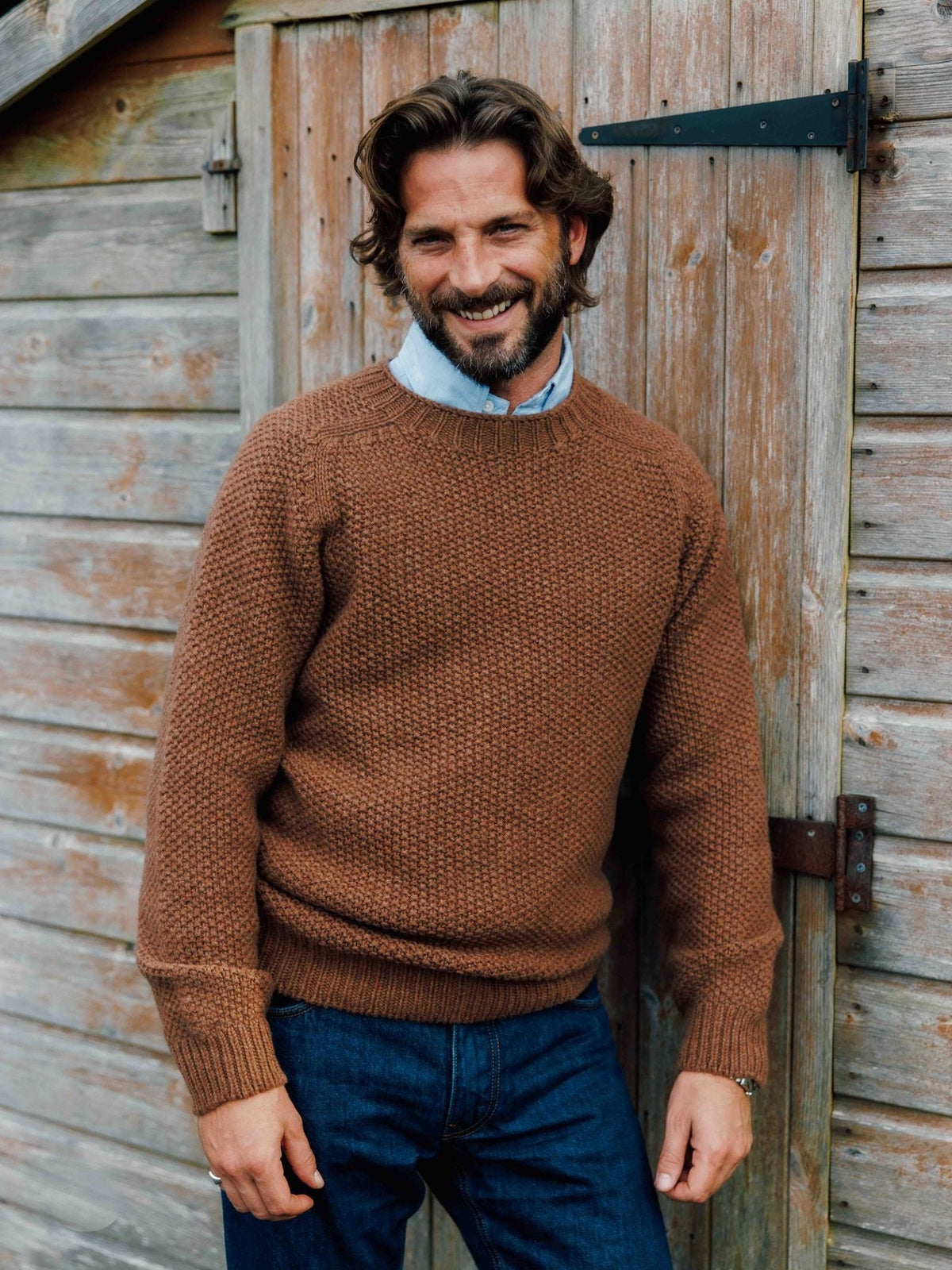 A smiling man wears a brown Campbell’s of Beauly Moss Stitch Crew Neck Jumper over a light blue collared shirt and jeans.