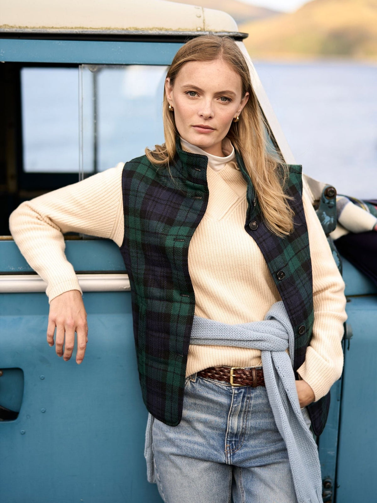 A woman wears a Campbell’s of Beauly Quilted Buttoned Tartan Gilet in Antique Black Watch over a cream ribbed sweater and blue jeans, leaning against the side of a blue vehicle.