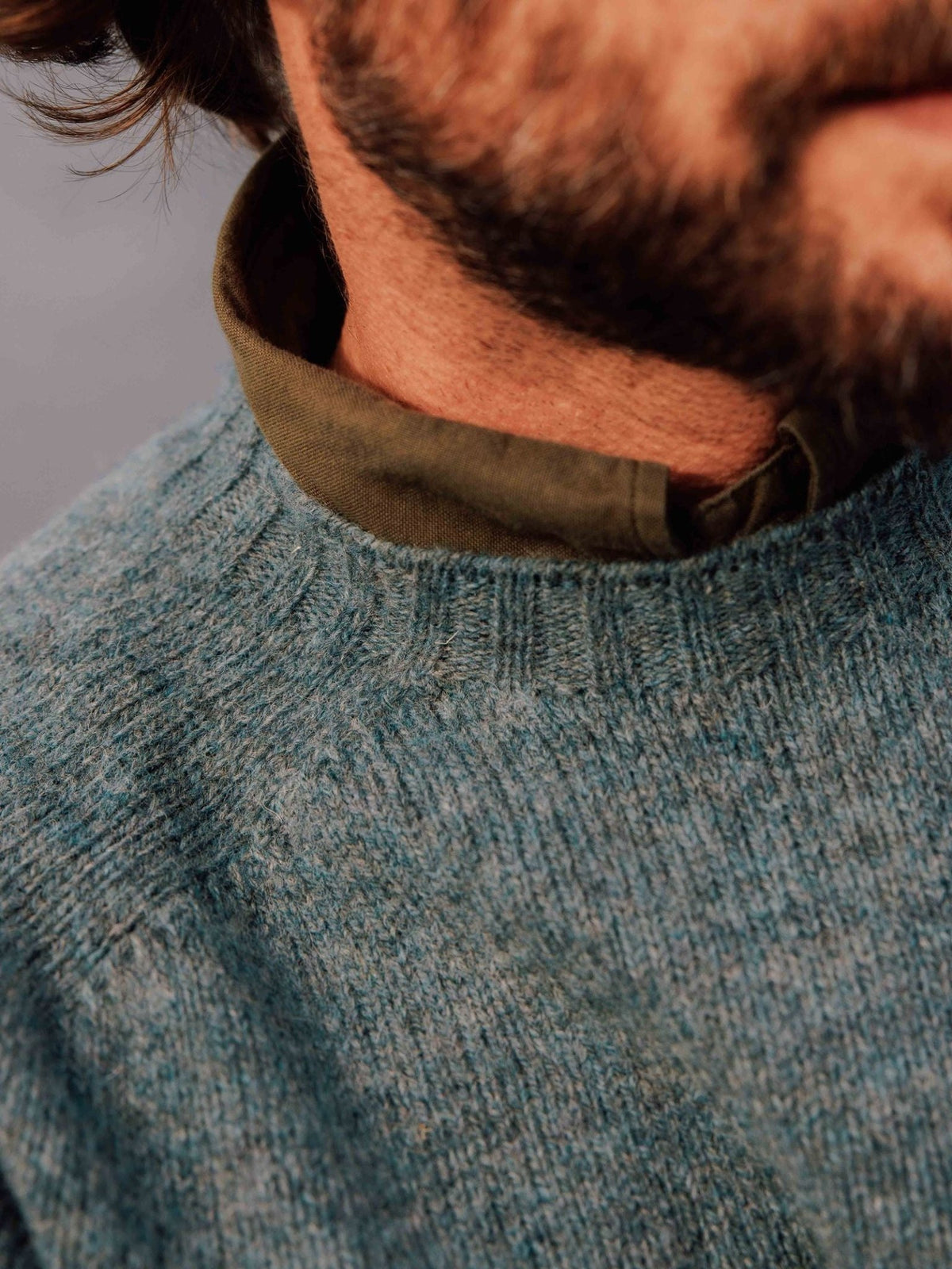 Close-up of a blue-grey Campbell’s of Beauly Shetland Jumper crewneck sweater worn over an olive green collared shirt.
