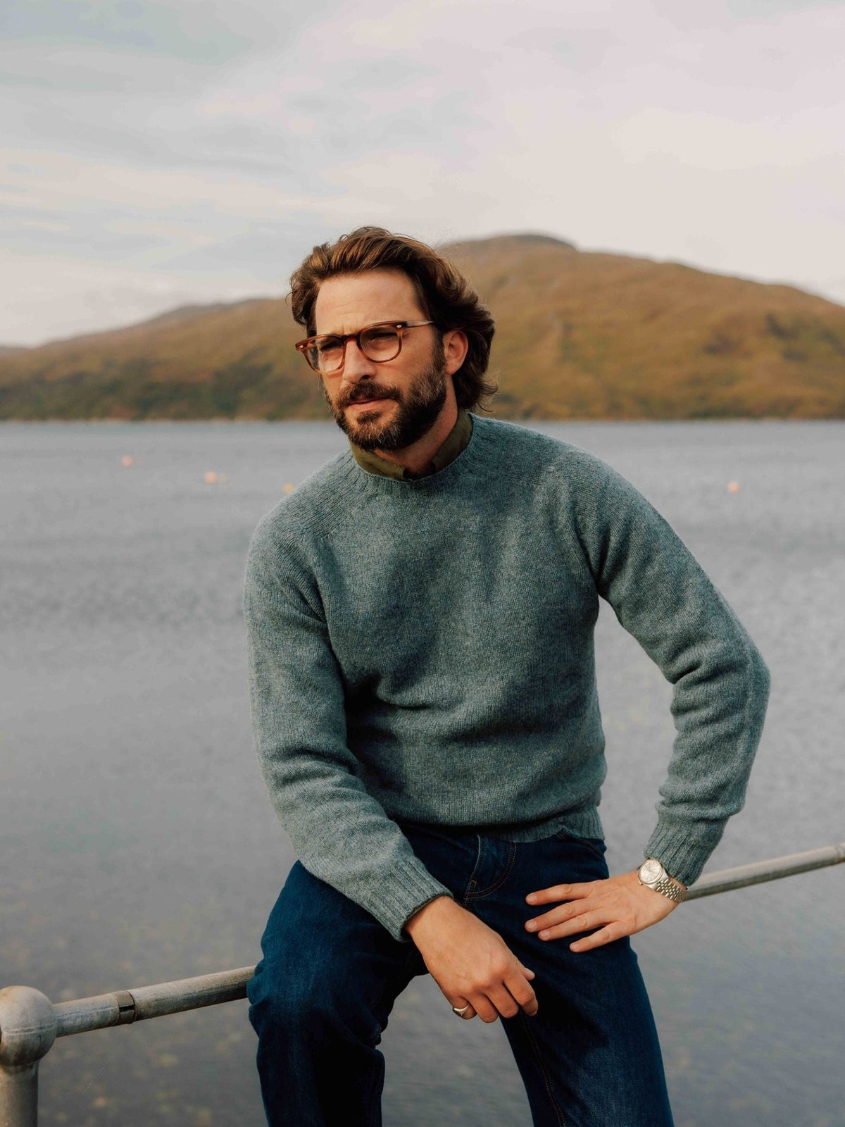 A man wearing a blue Shetland Jumper crew-neck sweater over an olive shirt, sitting on a metal railing by a lake with mountains in the background.