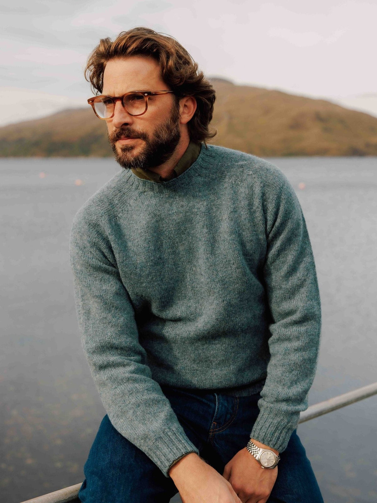 A man with a beard and glasses wears a light blue Campbell’s of Beauly Shetland Jumper and dark jeans while sitting by a lake.