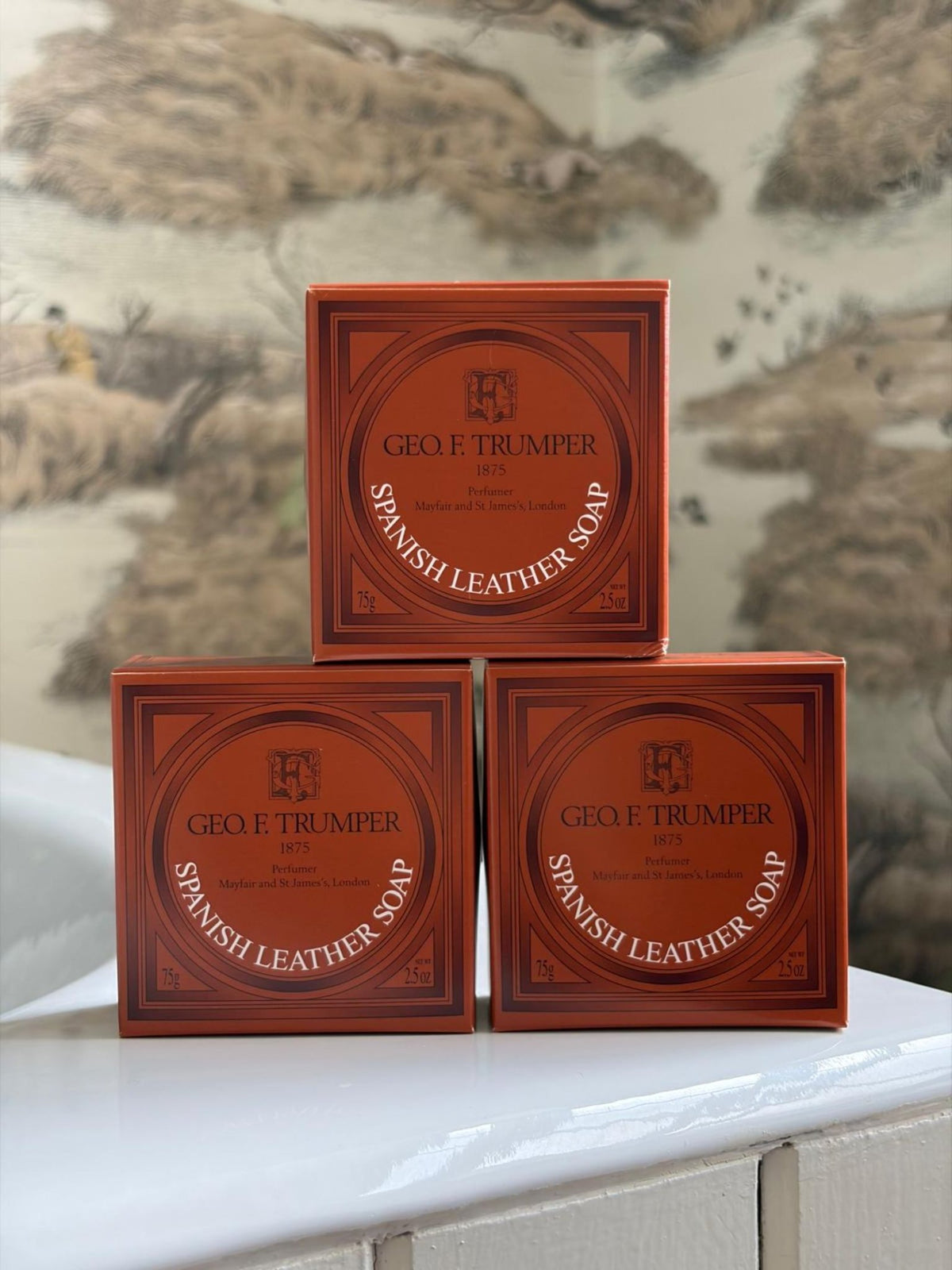 Three orange boxes of Geo. F. Trumper Spanish Leather Hand Soap, Set of 3 are stacked on a bathroom counter with vintage animal wallpaper—a refined addition to any guest bathroom.