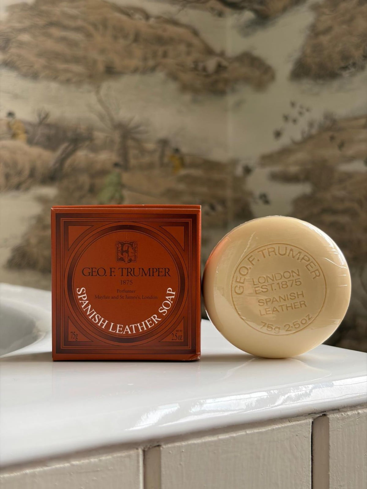 A box and bar of Geo. F. Trumper Spanish Leather Hand Soap, Set of 3 rest on white tiles with patterned wallpaper in the background—an elegant touch for guest bathrooms.