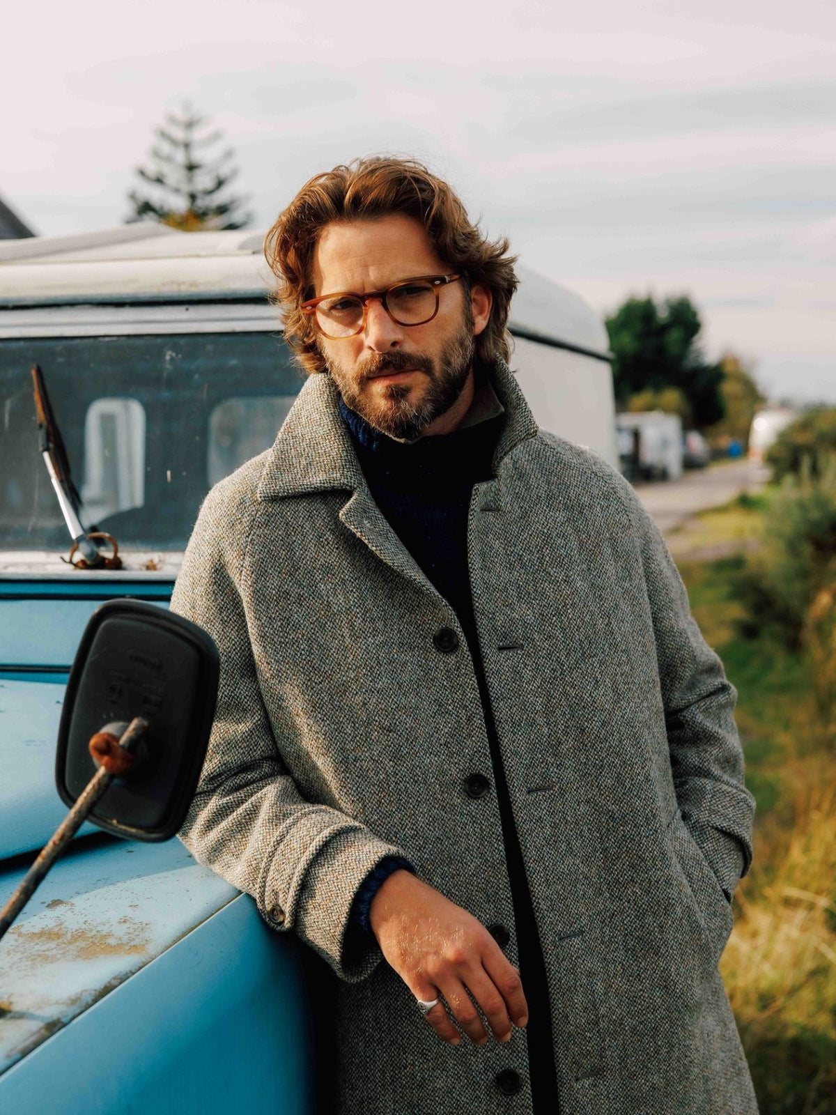 A man wearing a grey Harris Tweed Inverness Coat with a high collar leans against the side of a blue vehicle.