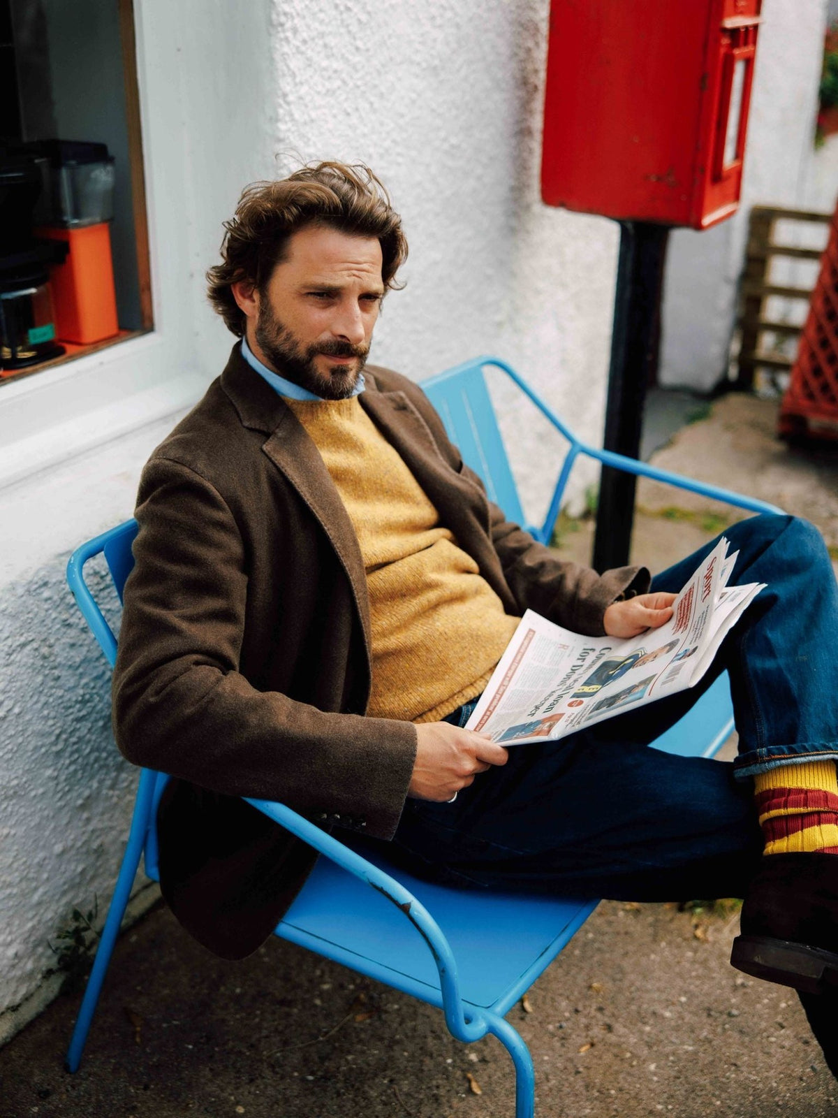 A bearded man wearing a Campbell’s of Beauly Washed Cashmere Jacket over a yellow sweater and blue jeans sits on a blue metal chair reading a newspaper.