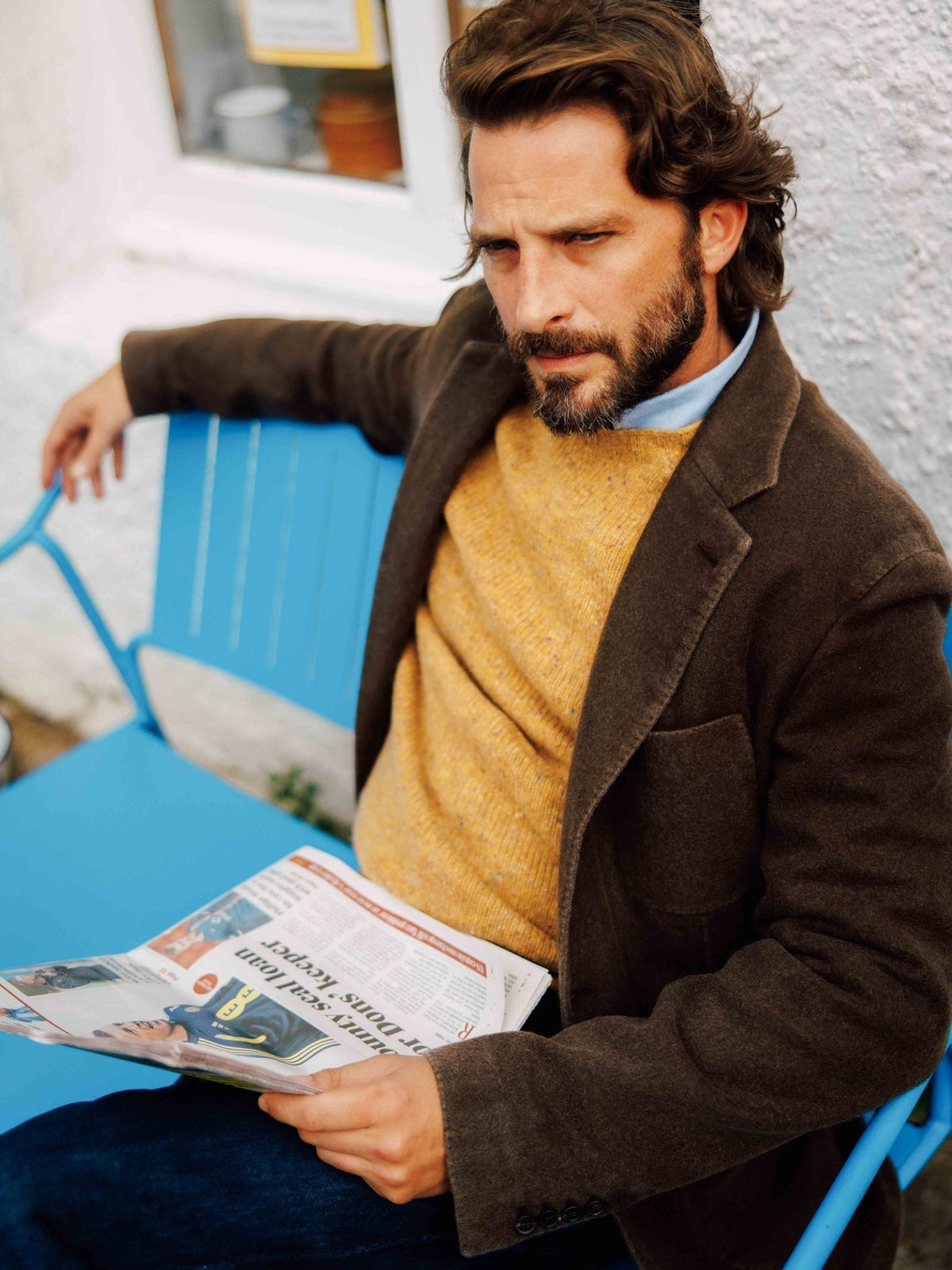 A bearded man wearing a Campbell’s of Beauly Washed Cashmere Jacket and a yellow sweater sits on a bright blue chair while reading a newspaper.