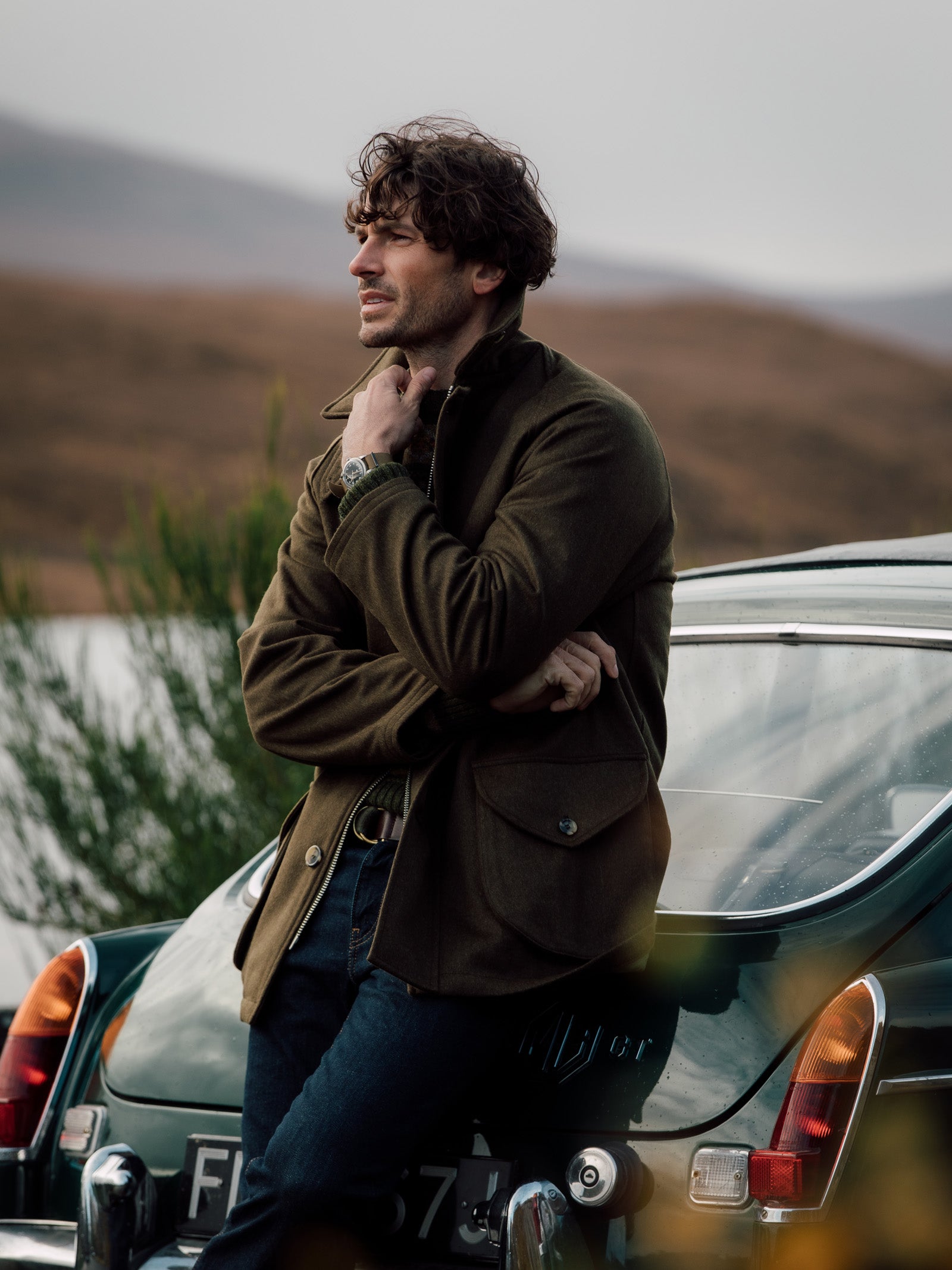 A man with curly brown hair in the Cannich Coat by Campbell’s of Beauly and jeans leans on a dark green vintage car, gazing thoughtfully across blurred hills beneath a cloudy sky, evoking timeless country pursuits.