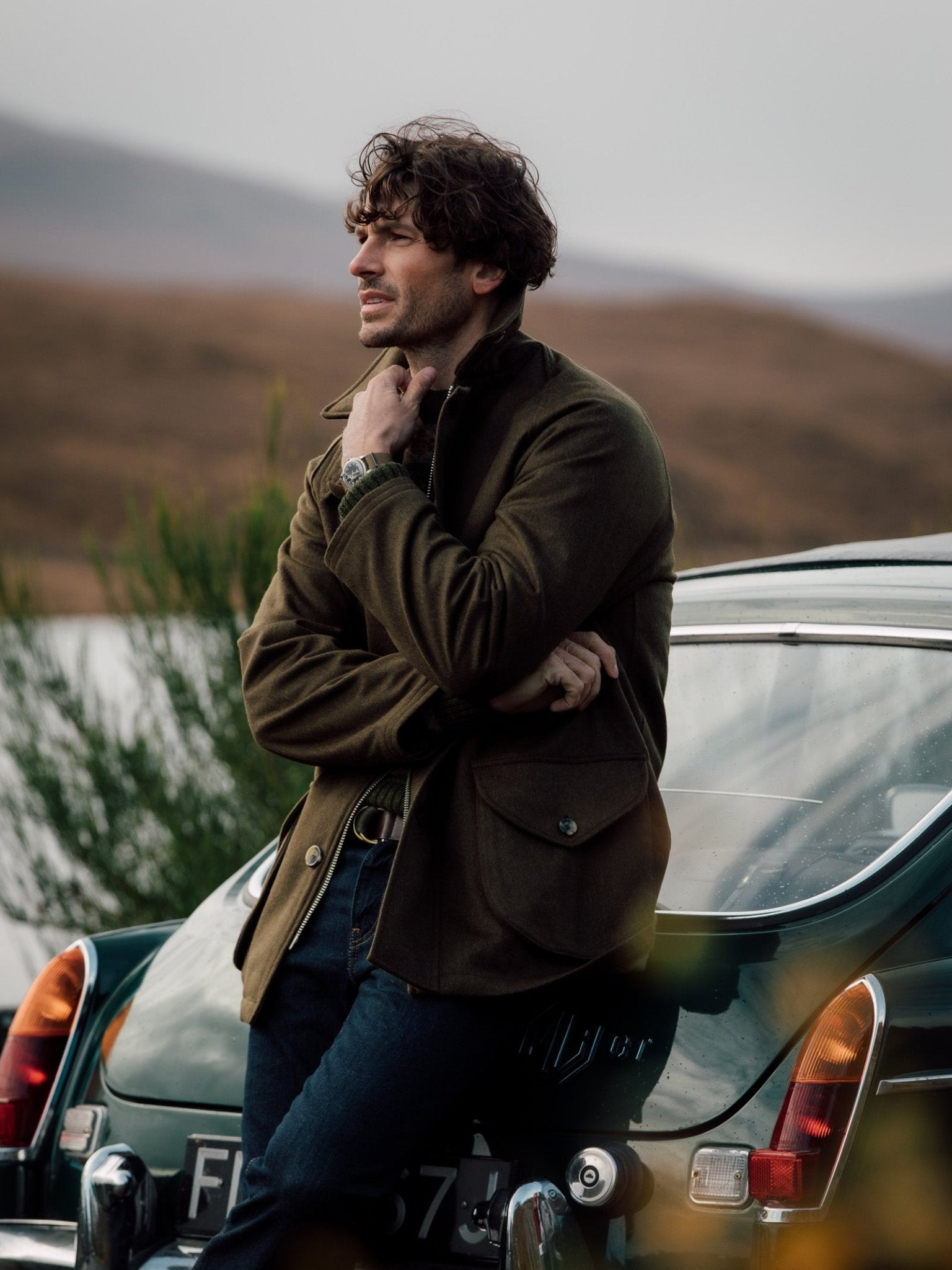 A man with wavy brown hair and stubble leans against a vintage dark green car outdoors, wearing the Cannich Coat by Campbells of Beauly over blue jeans as he gazes thoughtfully toward blurred hills and water, evoking classic country pursuits.