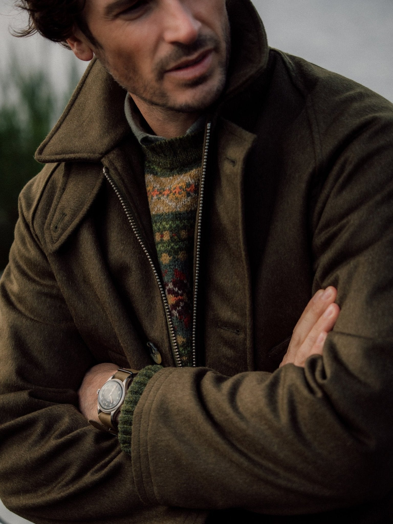 A man stands with arms crossed in a Campbells of Beauly Cannich Coat, collar up. He wears a patterned knit sweater and wristwatch, capturing classic country style against a blurred outdoor backdrop, his face partially visible and looking downward.