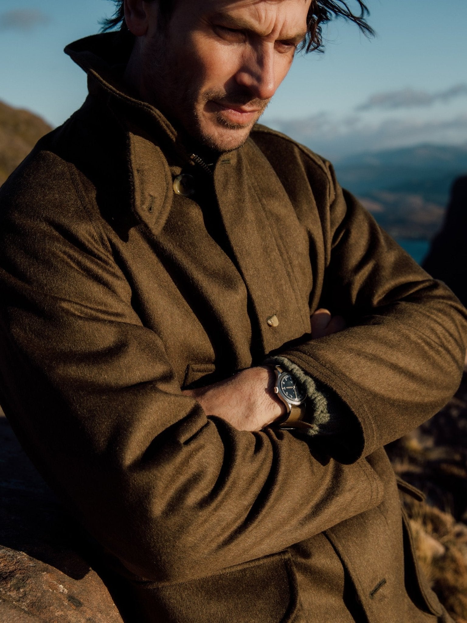 A man outdoors stands with arms crossed, wearing the Cannich Coat by Campbells of Beauly, crafted from Austrian wool. His brown hair is windswept as he gazes downward against rocky hills and a cloudy sky in soft sunlight.