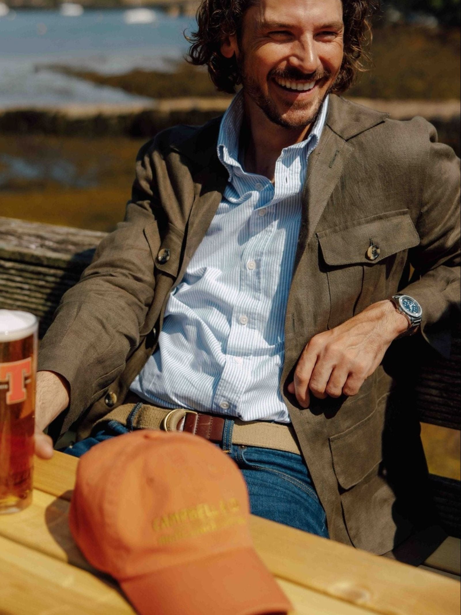 A smiling man in a brown blazer and blue shirt, wearing the Campbell's of Beauly Canvas D-Loop Belt, sits outdoors with a glass of beer. An orange baseball cap is on the wooden table, with water and greenery blurred behind him.