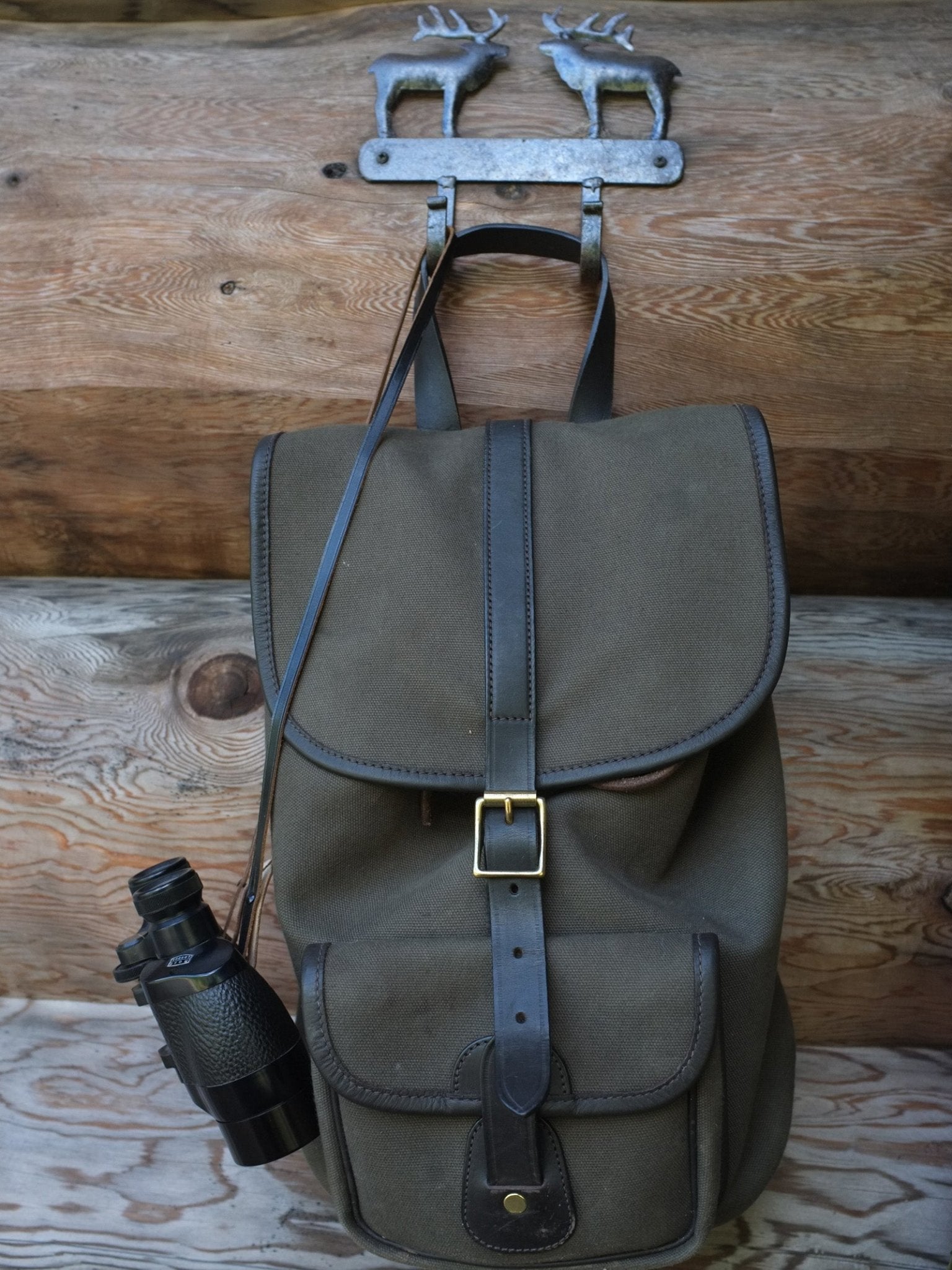 A Campbells of Beauly Canvas Rucksack with dark brown leather straps hangs on a wooden wall beneath a metal deer-shaped hook, with black binoculars attached to its side.