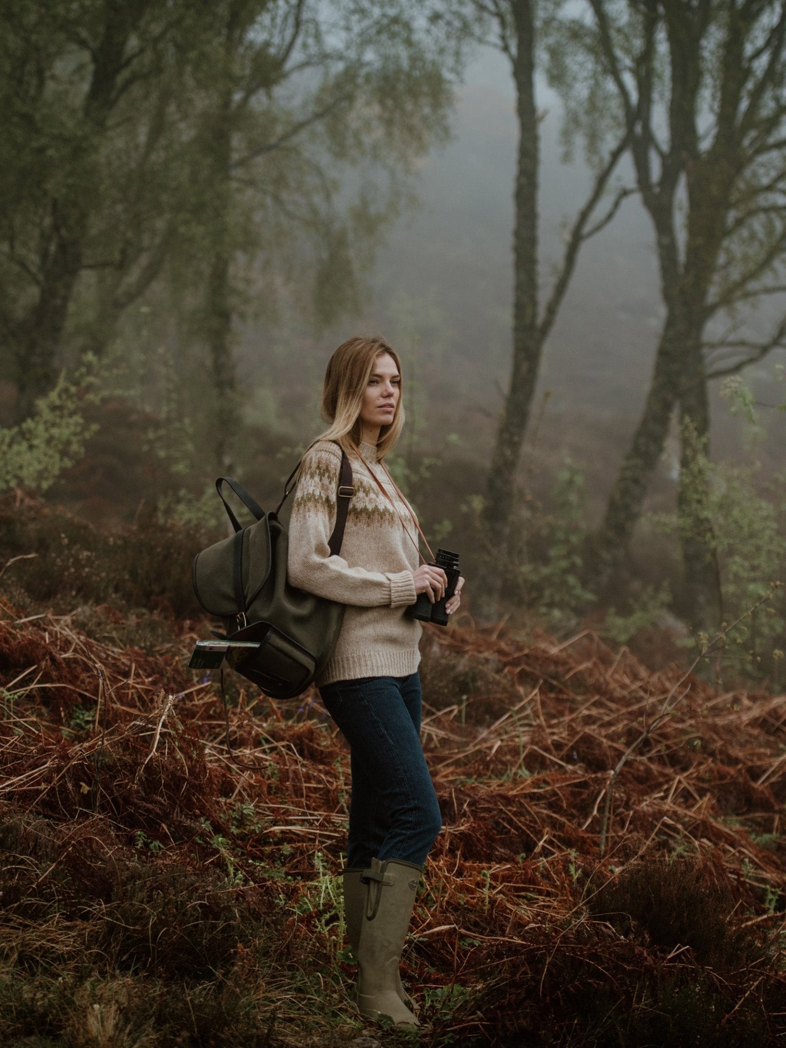 A woman stands in a misty forest, wearing a sweater, jeans, and boots. She carries the Campbells of Beauly Canvas Rucksack and holds binoculars amid brown ferns and tall trees.