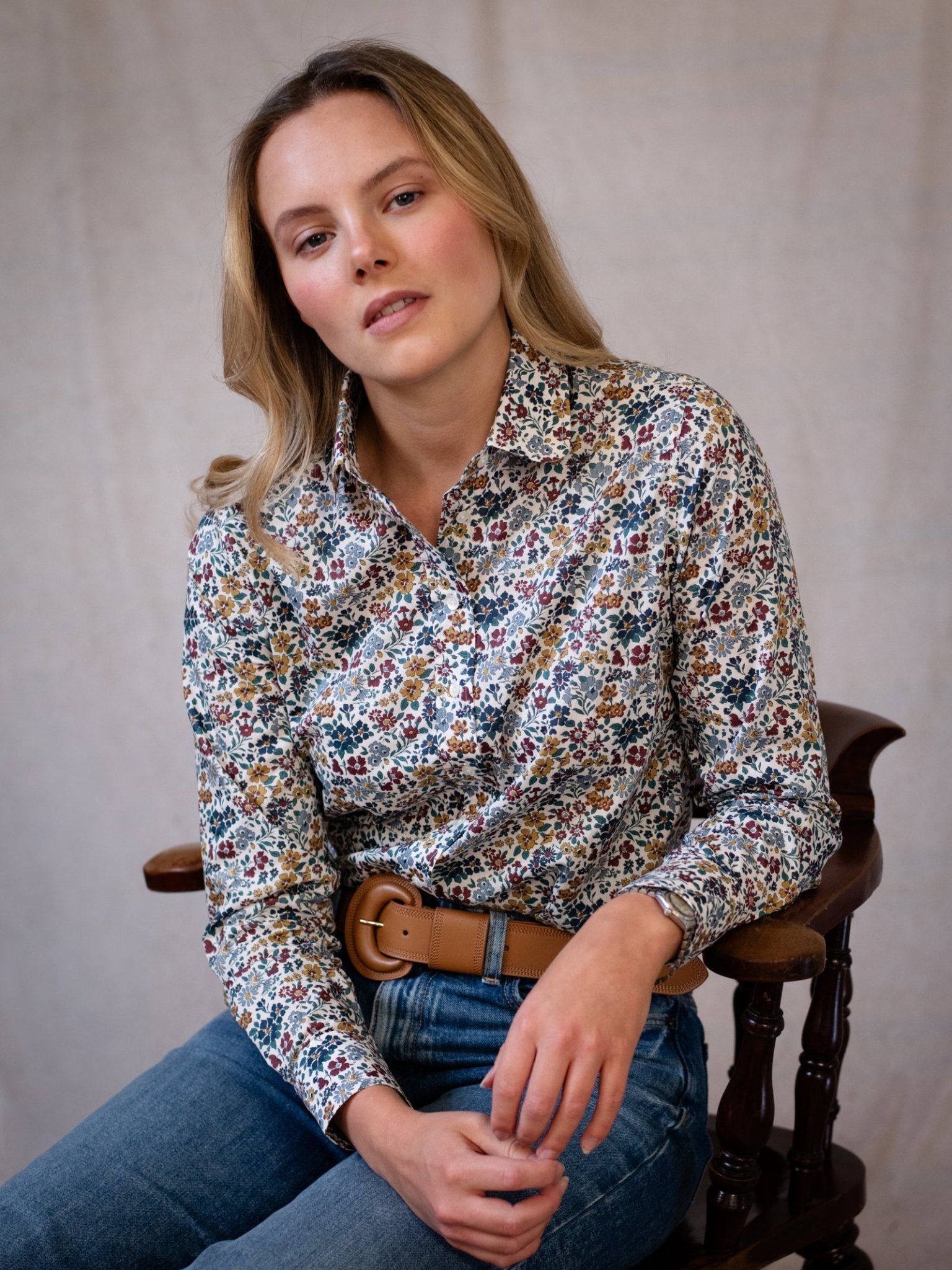 A woman with long blonde hair sits on a wooden chair, wearing a floral shirt, blue jeans, and the Caramel Leather Belt by Campbell's of Beauly. Set against a light background, she gazes at the camera with a relaxed expression.