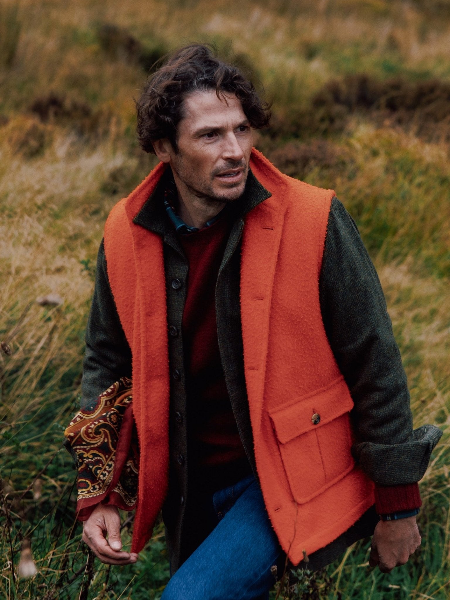 A man with wavy brown hair stands in a grassy field, wearing a dark green coat over a bright orange Campbells of Beauly Casetino Fleece Gilet, a red sweater, and blue jeans. He holds patterned fabric under his arm and looks to the side.